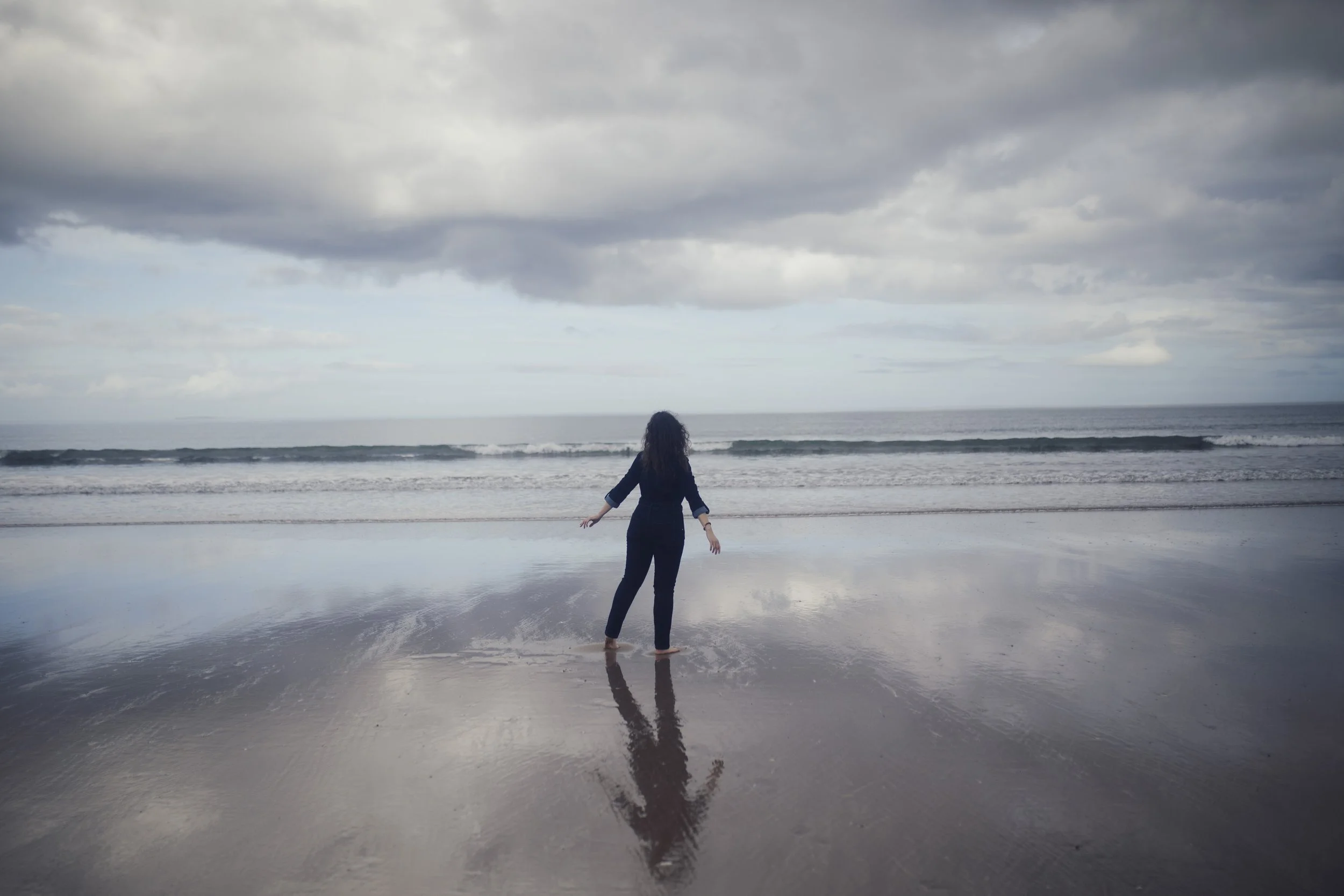 A woman in dark clothing standing on a wet beach looking toward the ocean, with clouds above and her reflection visible on the water.