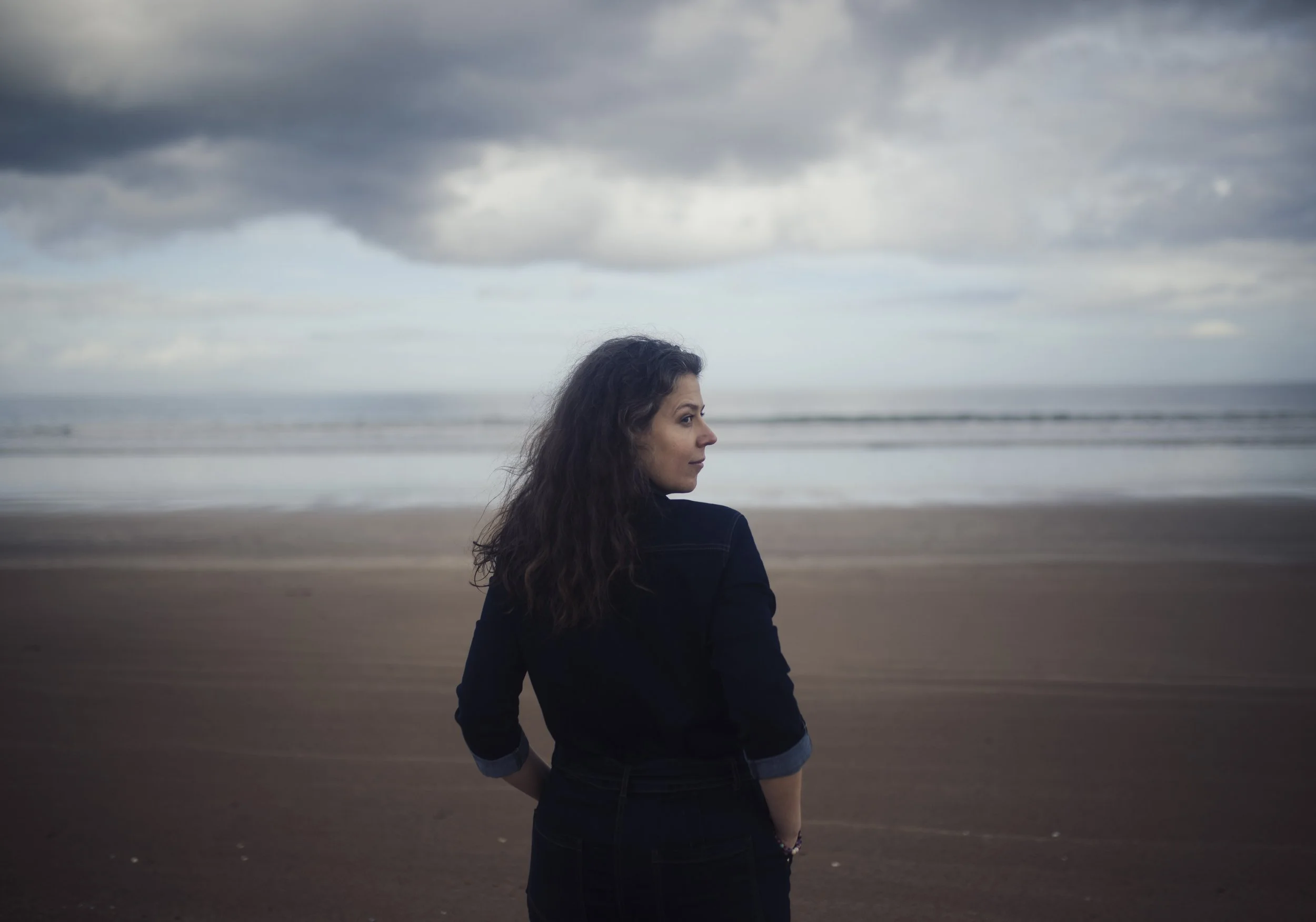 A woman with dark, wavy hair stands on a beach facing sideways with her hands on her hips, looking towards the ocean under a cloudy sky.