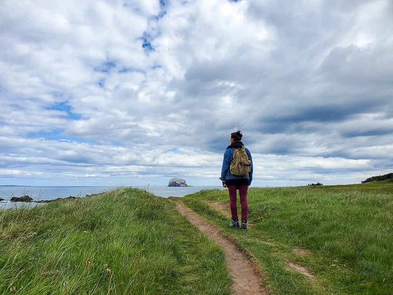 A woman walking on a grassy trail along the coast, with a backpack, looking towards a large rock island in the ocean under a cloudy sky.