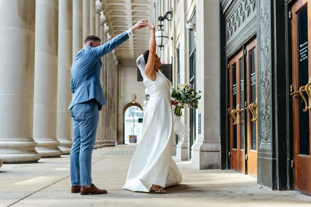 A bride and groom dancing outside a building with tall columns, the bride holding a bouquet.
