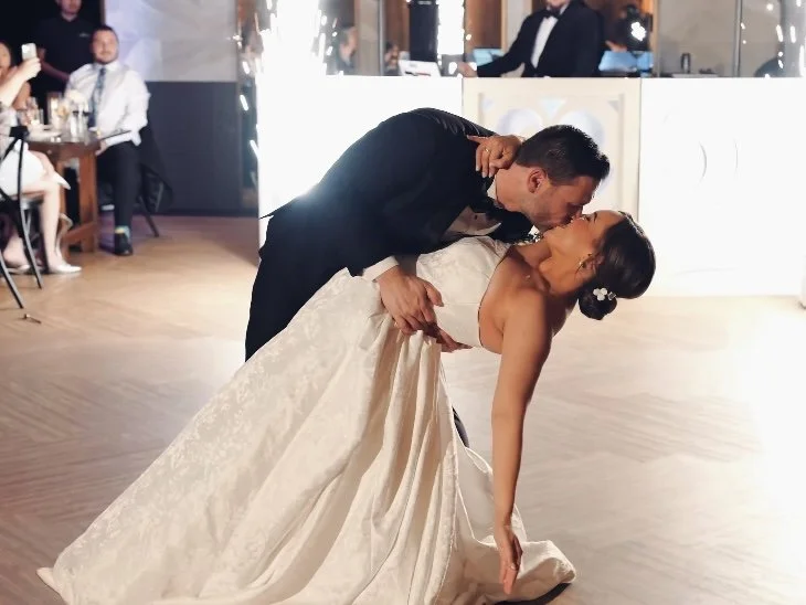 A couple dancing at a wedding reception, with the groom dipping the bride in a ballroom dance move as they share a kiss. The bride is wearing a white gown, and the groom is in a black suit. Guests are seated in the background.