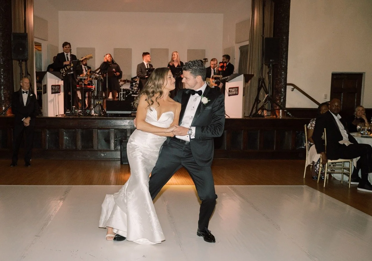 Bride and groom dancing at a wedding reception, with a live band performing in the background and guests watching.