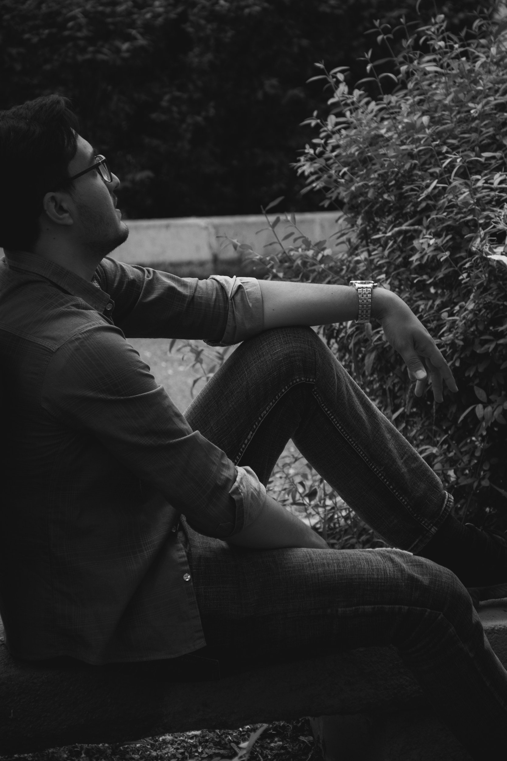 A man sitting on a park bench, looking at the greenery in front of him, in black and white.