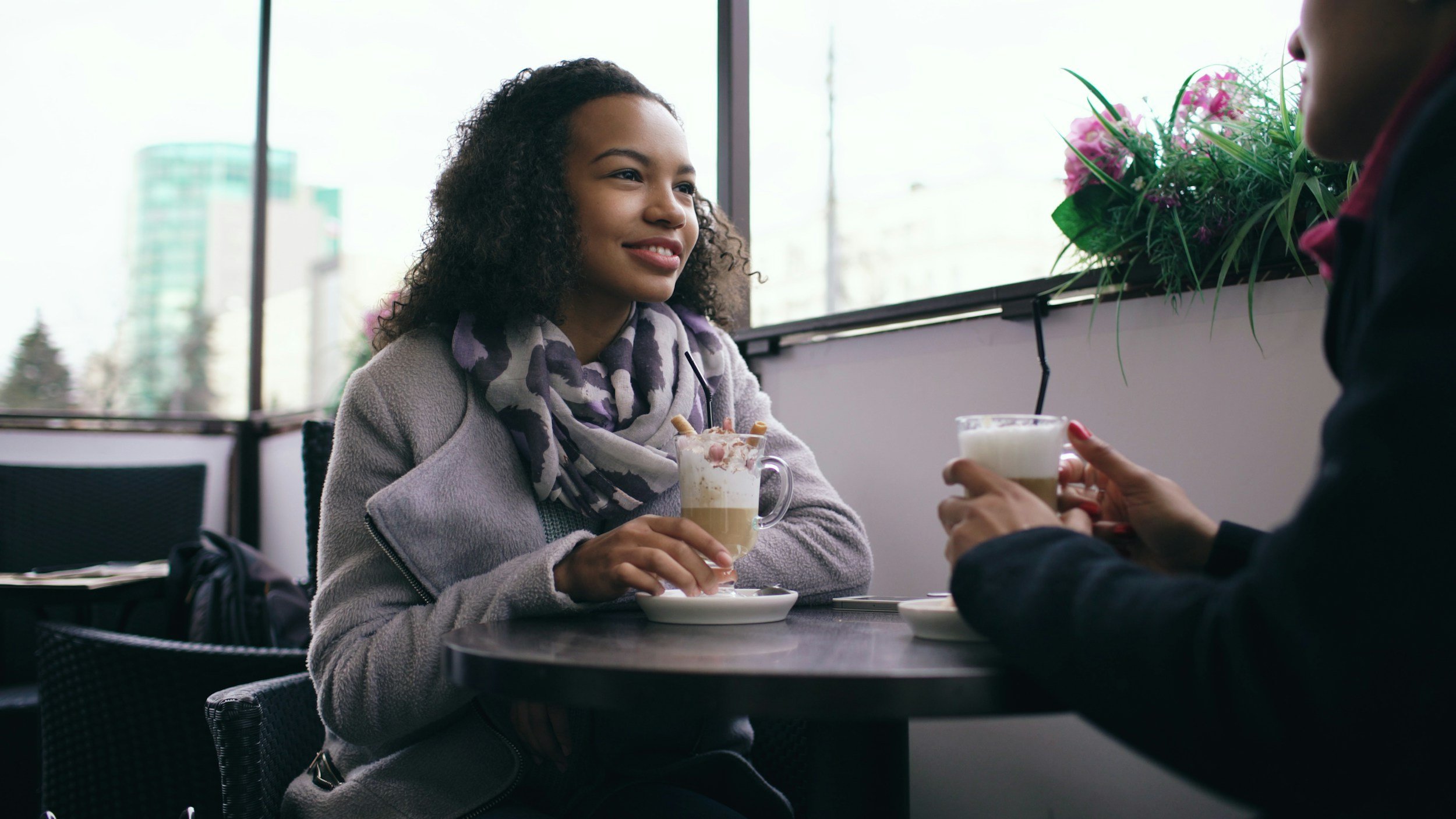 Two women sitting at a table in a cafe, enjoying coffee drinks with whipped cream in glass mugs. One woman is smiling and wearing a gray sweater and a purple patterned scarf, while the other woman, partially visible, holds her coffee mug.