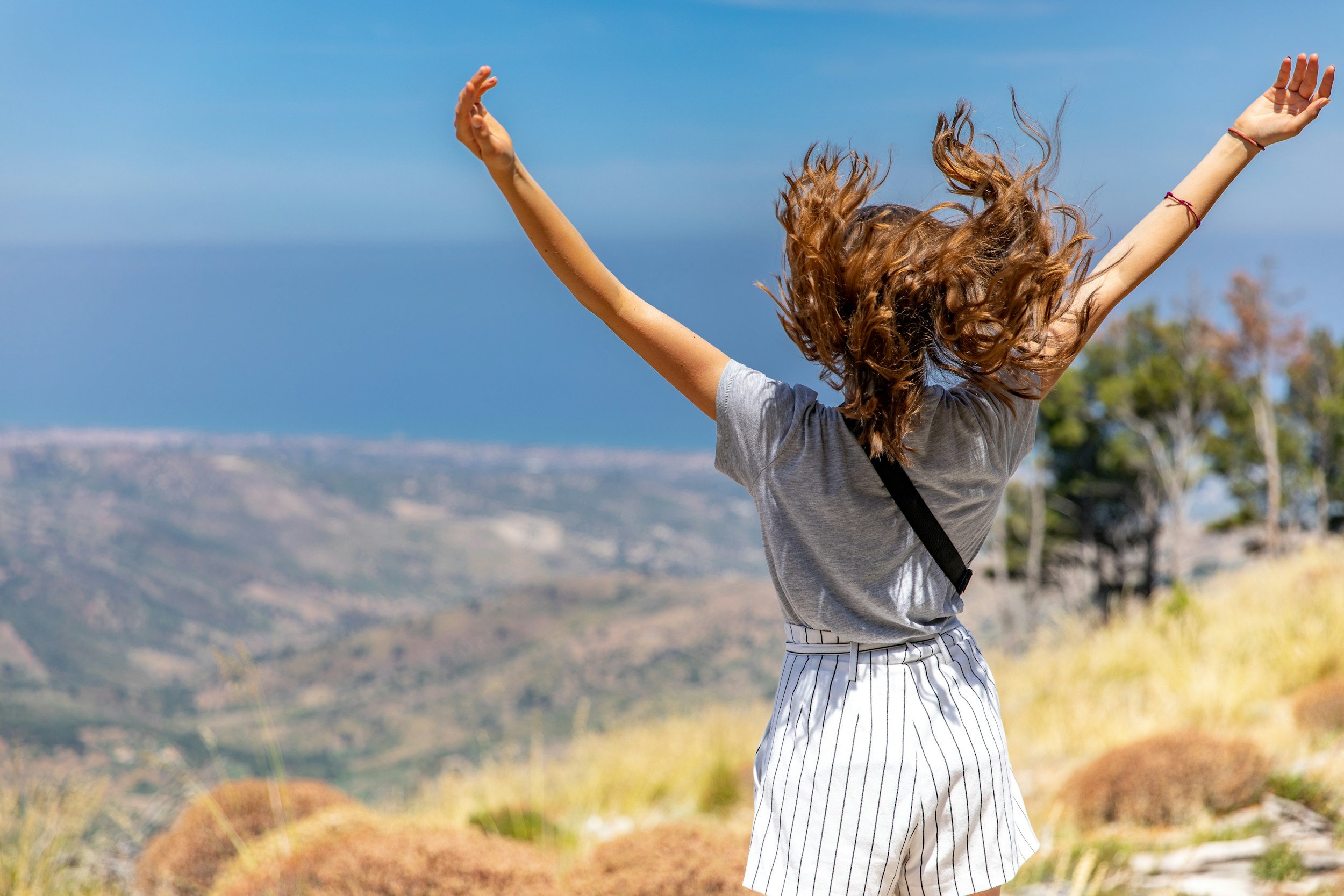 A woman with curly hair standing on a hilltop with her arms raised, overlooking a landscape with hills and the ocean under a clear blue sky.