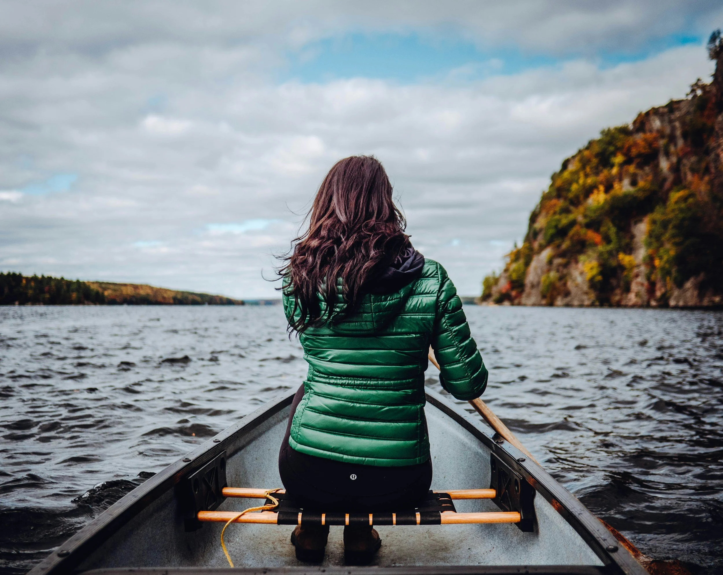 A woman with long dark hair sitting in a canoe on a river, facing away, wearing a green puffer jacket, surrounded by water with hills and trees in the distance.