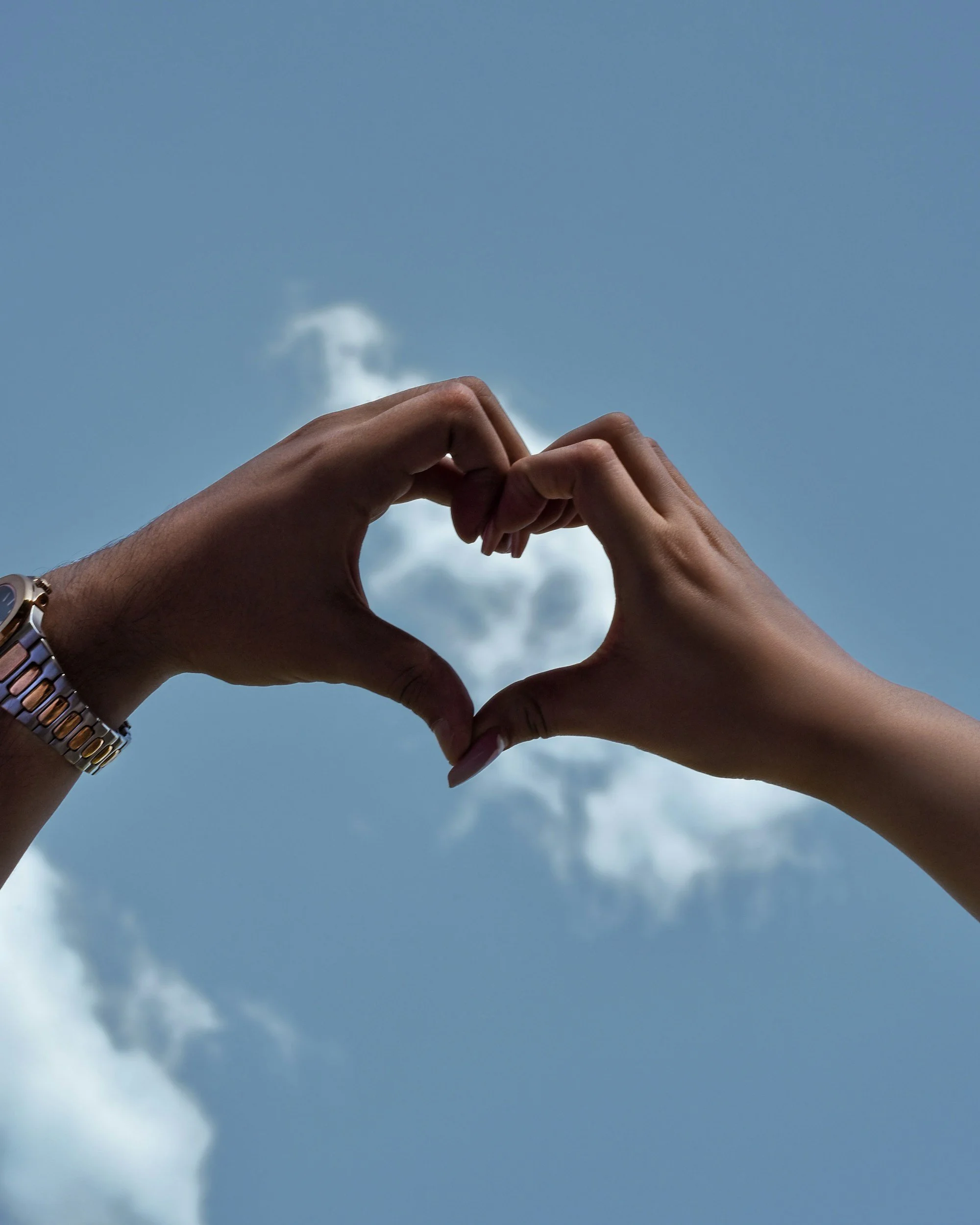 Two hands form a heart shape against a blue sky with white clouds.