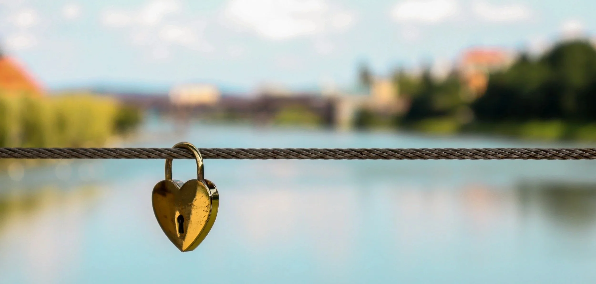 A close-up of a golden heart-shaped lock on a wire with a blurred background of a river, bridge, trees, and buildings.