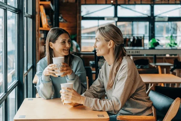 Two women sitting at a table in a coffee shop, smiling and enjoying their drinks while talking.