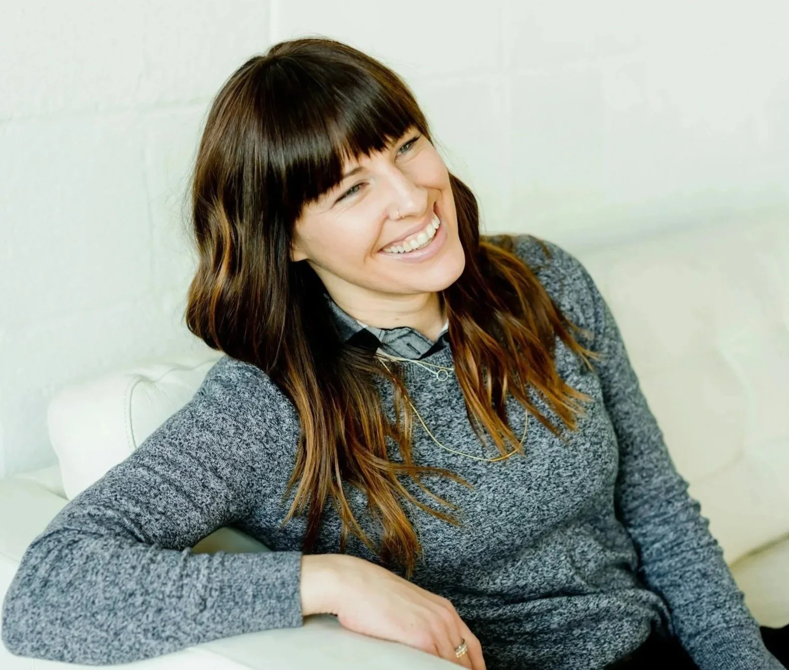 A woman with brown hair and bangs, smiling and sitting on a white couch.