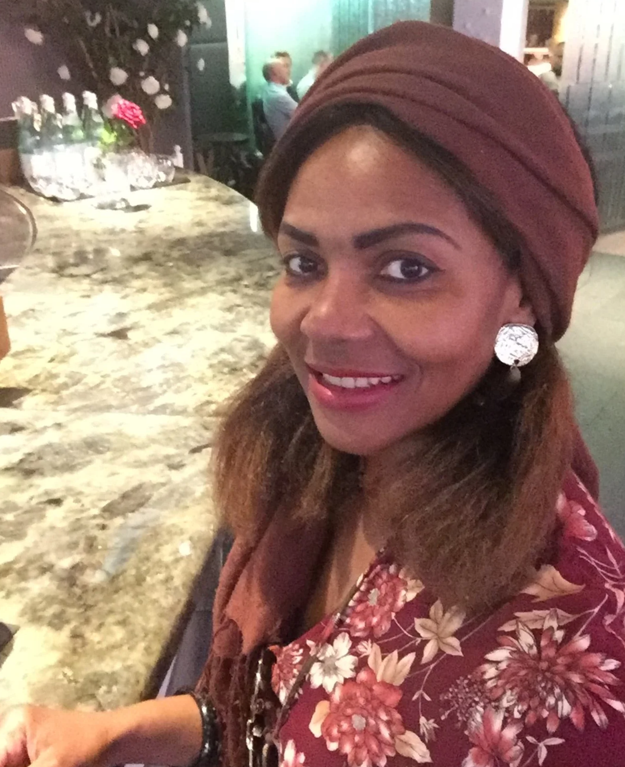 A woman with a brown headwrap, floral-patterned blouse, and large earrings sitting at a marble counter in a restaurant.