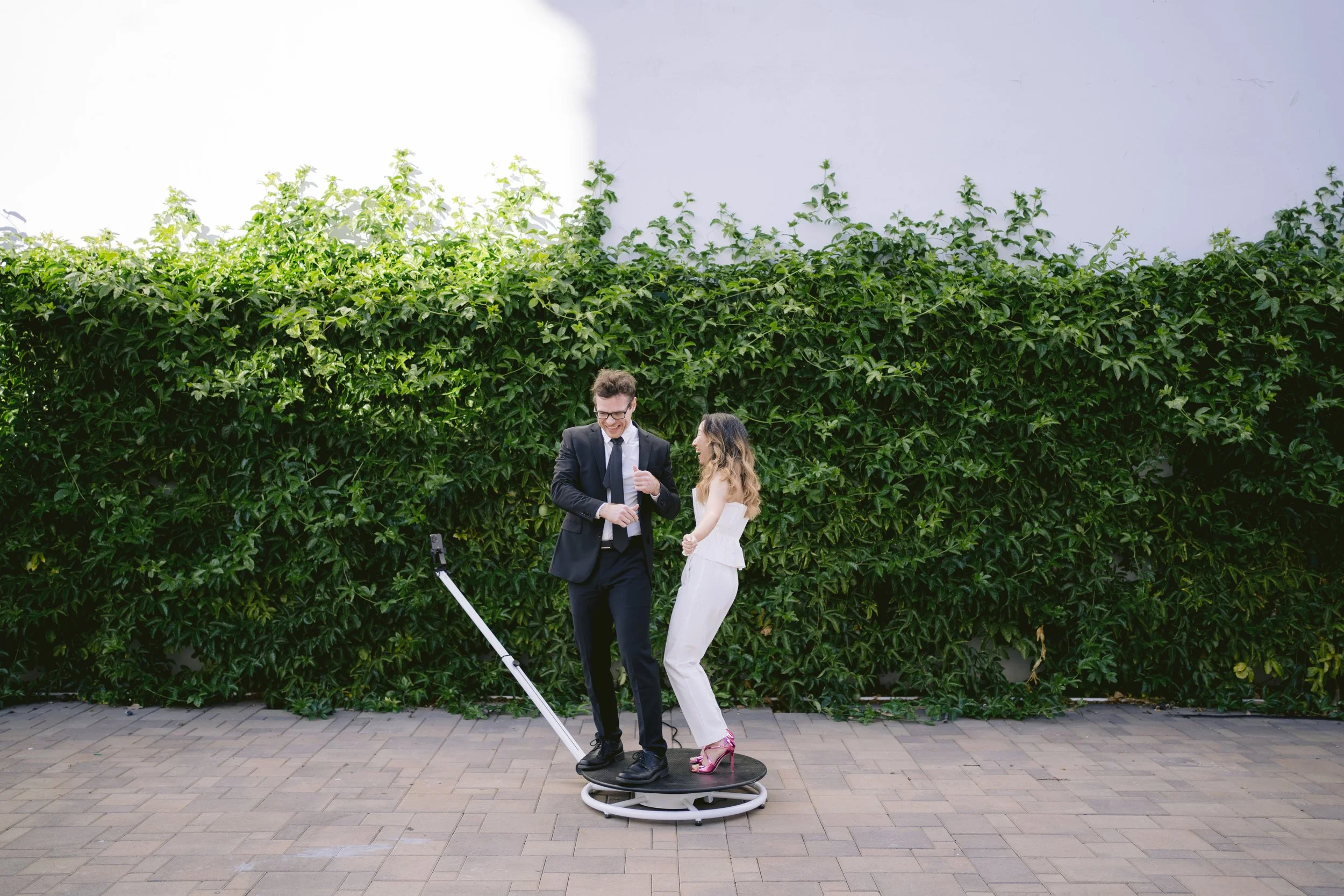 A man in a black suit and a woman in a white outfit dancing on a 360 photo booth outdoors against a green hedge background.