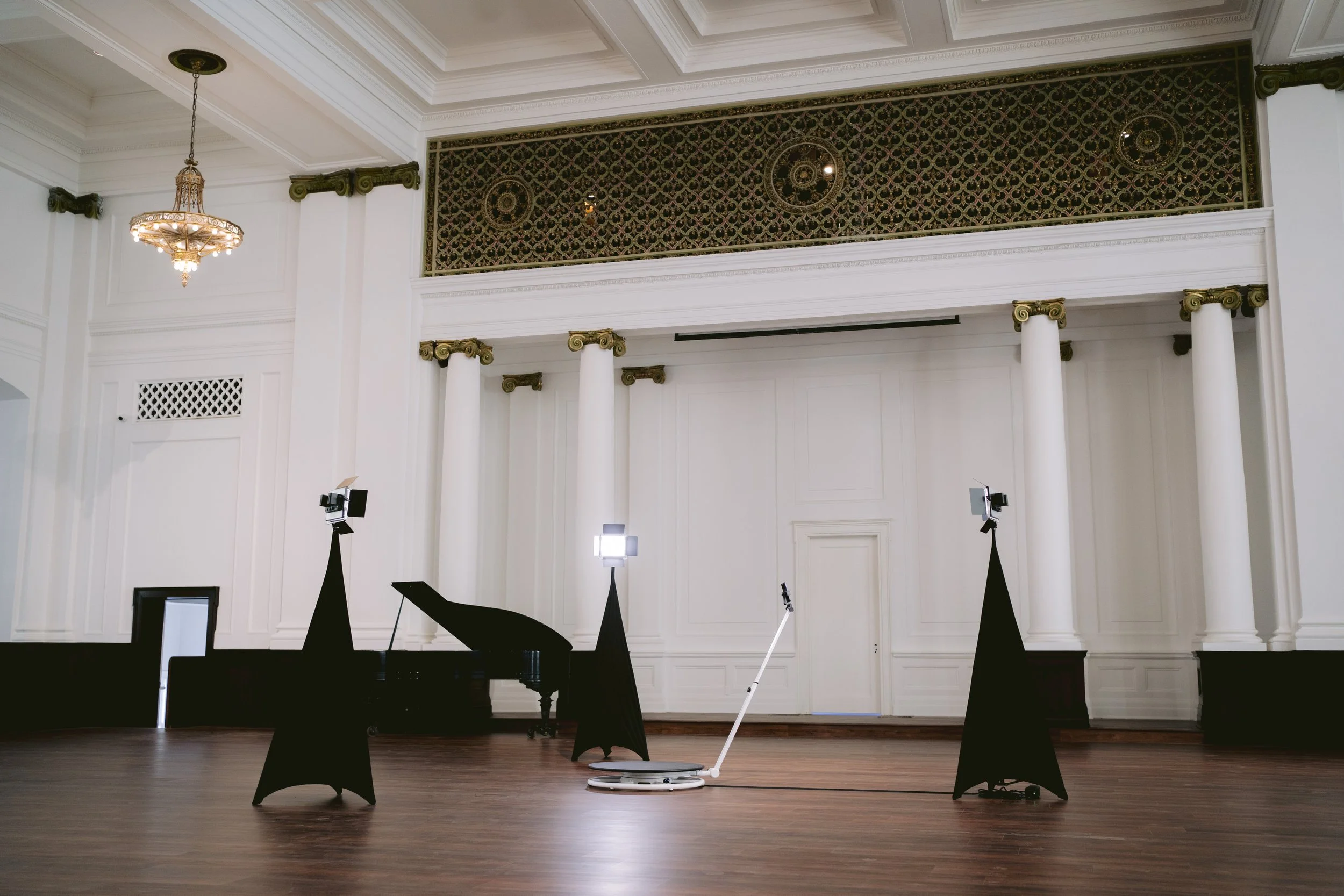 Empty stage with a black grand piano, lighting equipment, and a white 360 photo booth in a large, ornate hall with white walls, columns, and decorative gold accents.