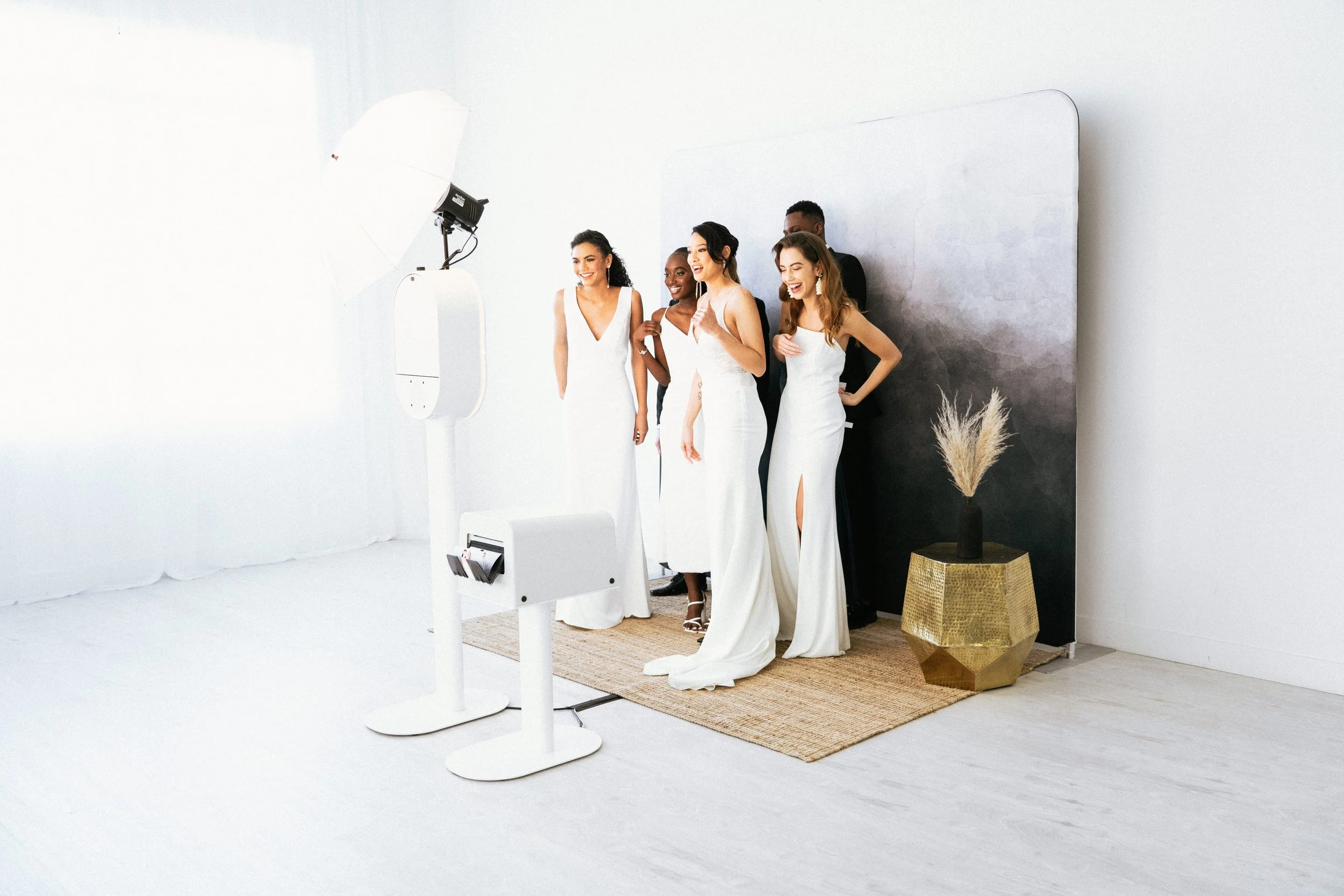 Group of five women in white dresses posing and smiling at a photo booth in a bright, white studio with a black background and gold vase with dried plants.
