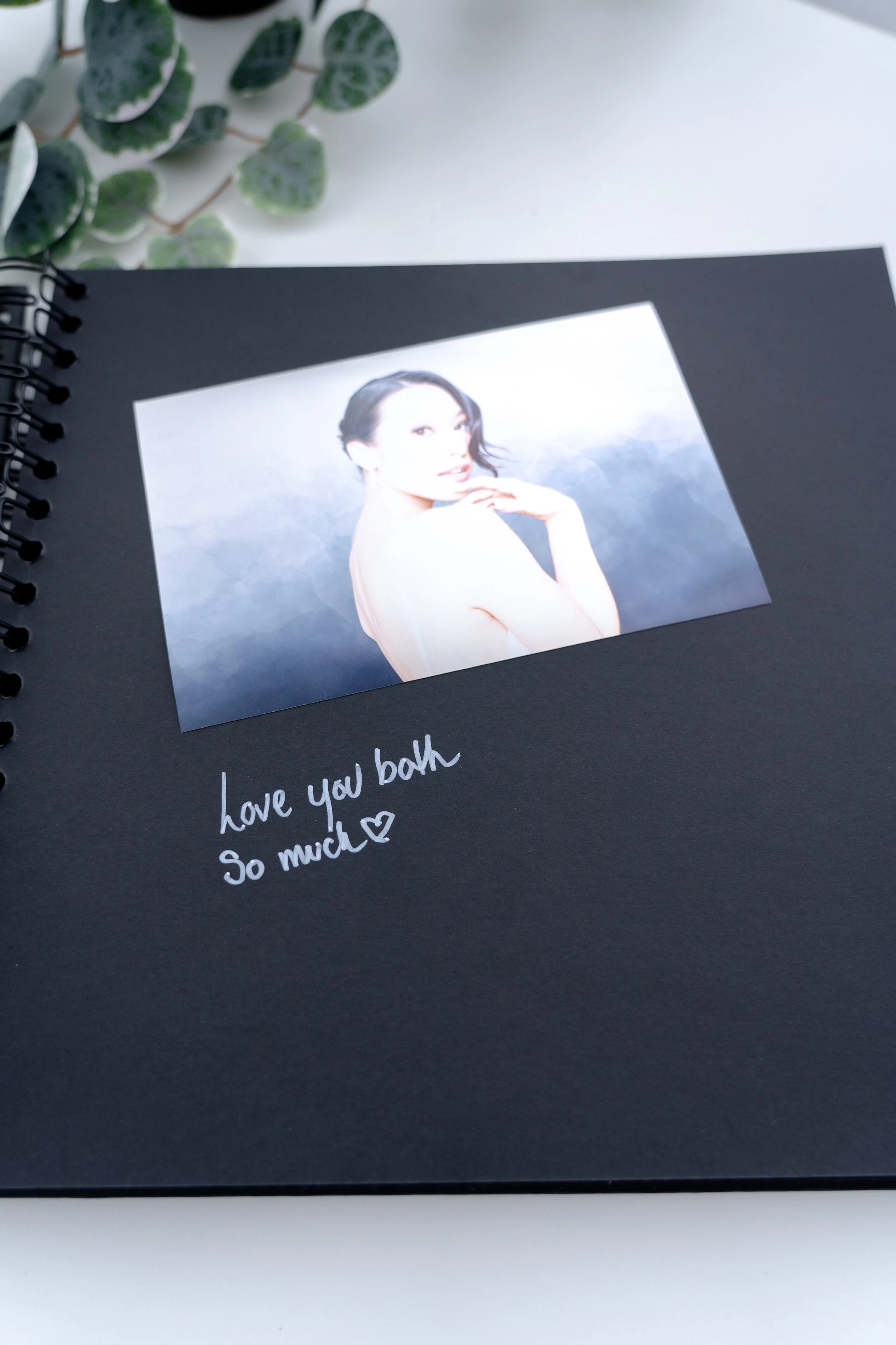 A black scrapbook or photo album with a photo of a woman on a cloudy background, and handwritten text that says 'love you both so much' with a heart.