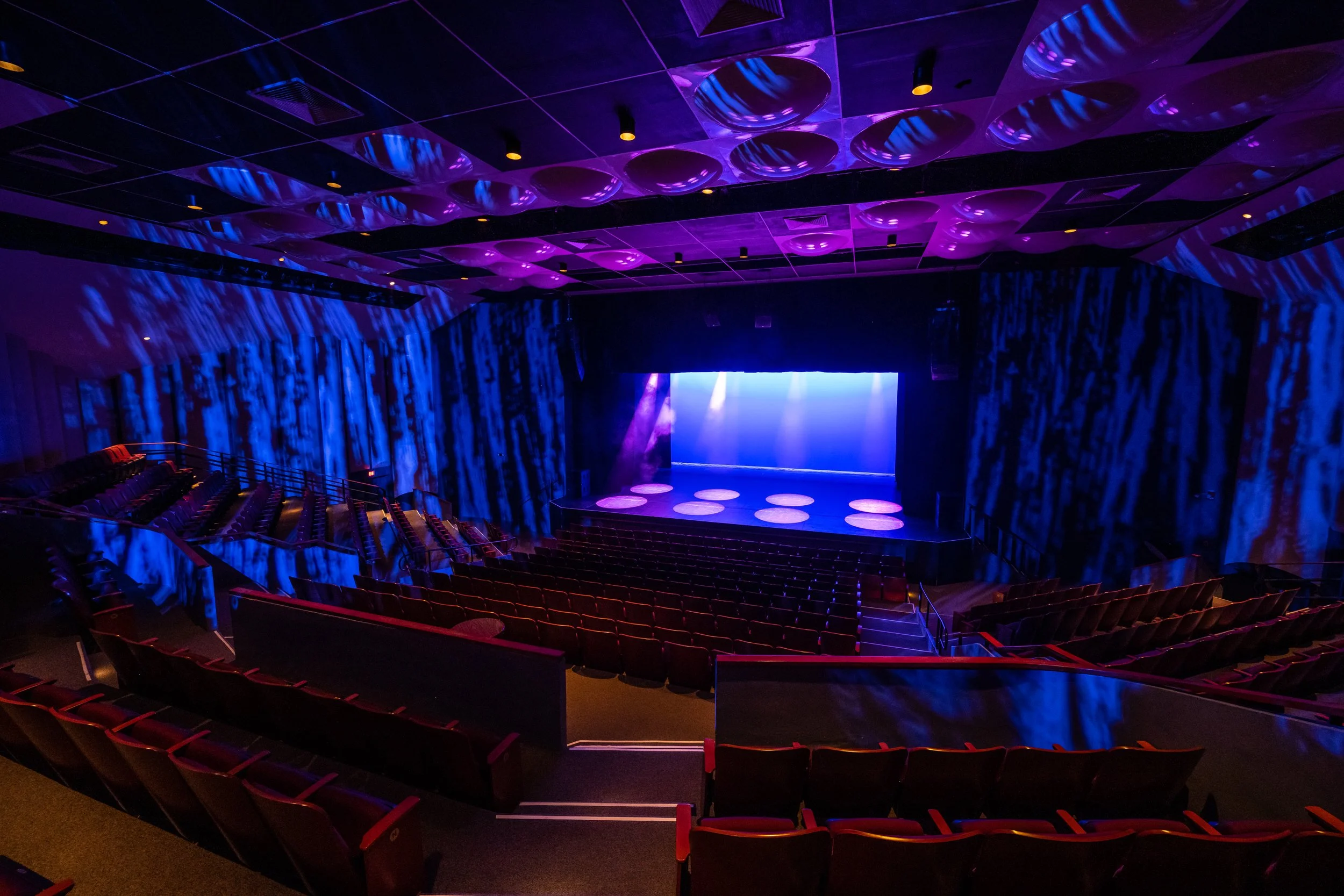 An empty theater auditorium with blue and purple lighting, a stage with six circular platforms, and rows of red seats.