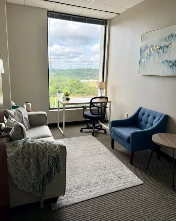 A modern office room with a large window showing a green landscape. The room has a white couch with pillows, a blue armchair, a round wooden side table, a white desk with a black office chair, and a large abstract painting on the wall. A beige rug covers part of the carpeted floor.