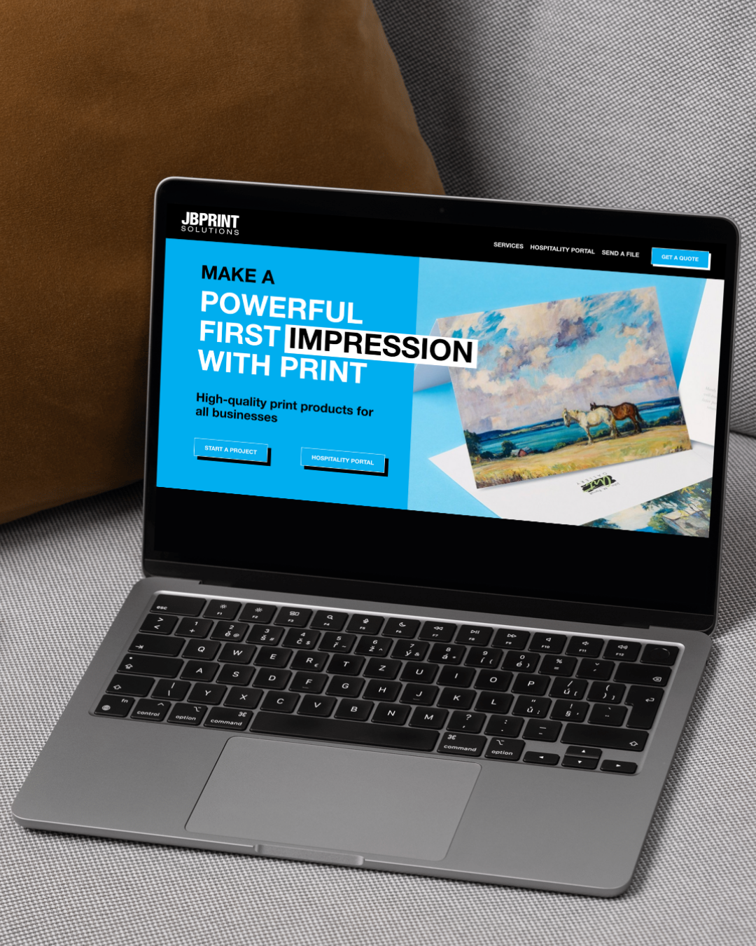 Laptop displaying a JB Print Solutions website with a focus on high-quality print products. The screen features a promotional message and images of printed materials. The laptop is placed on a gray fabric surface with a brown cushion nearby.