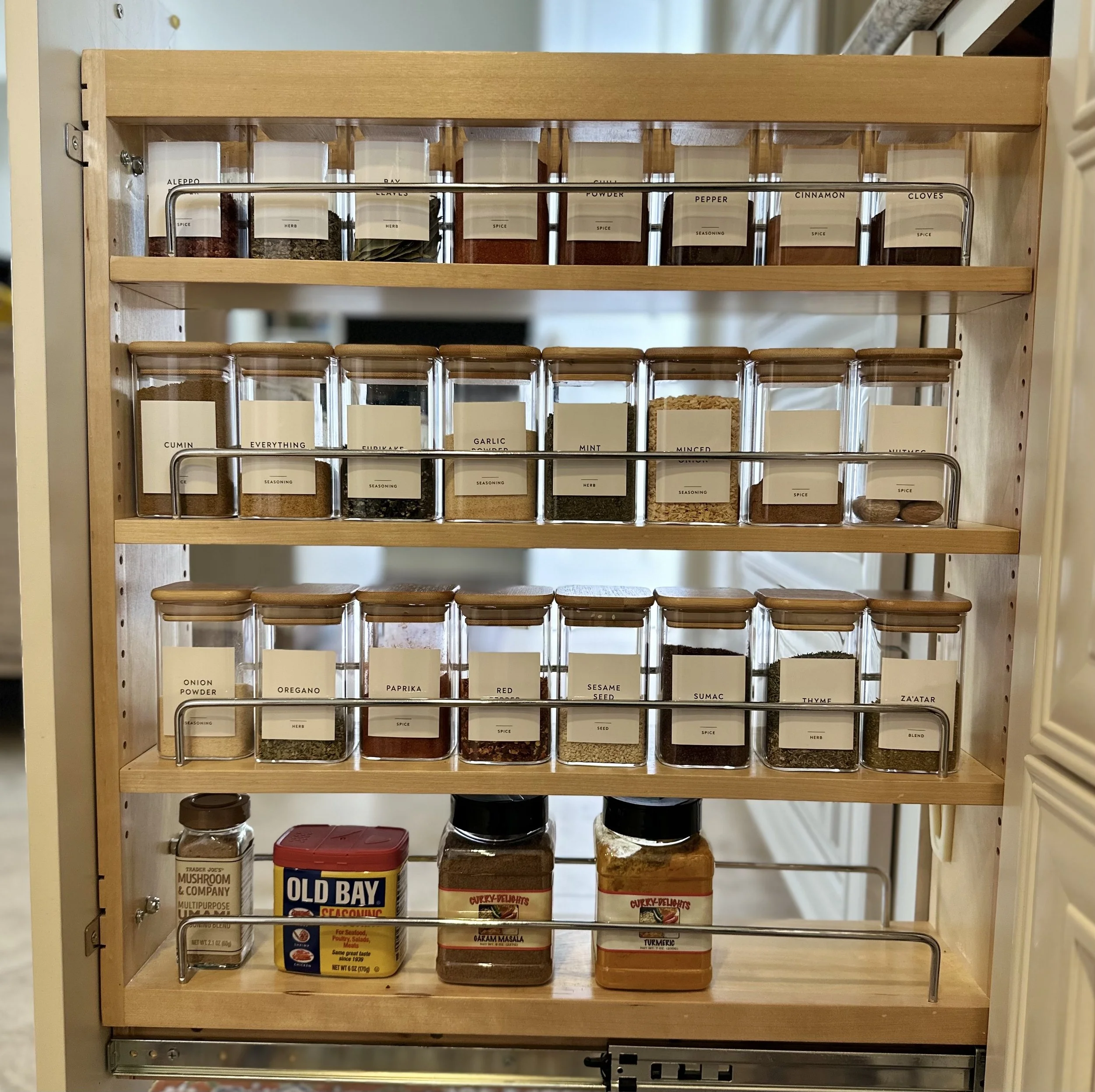 Organized wooden spice rack with labeled glass jars of spices including cumin, garlic, mint, oregano, paprika, and turmeric. Bottom shelf holds larger containers like mushroom seasoning and curry powder. Styled by Tidy Doon, Los Angeles.