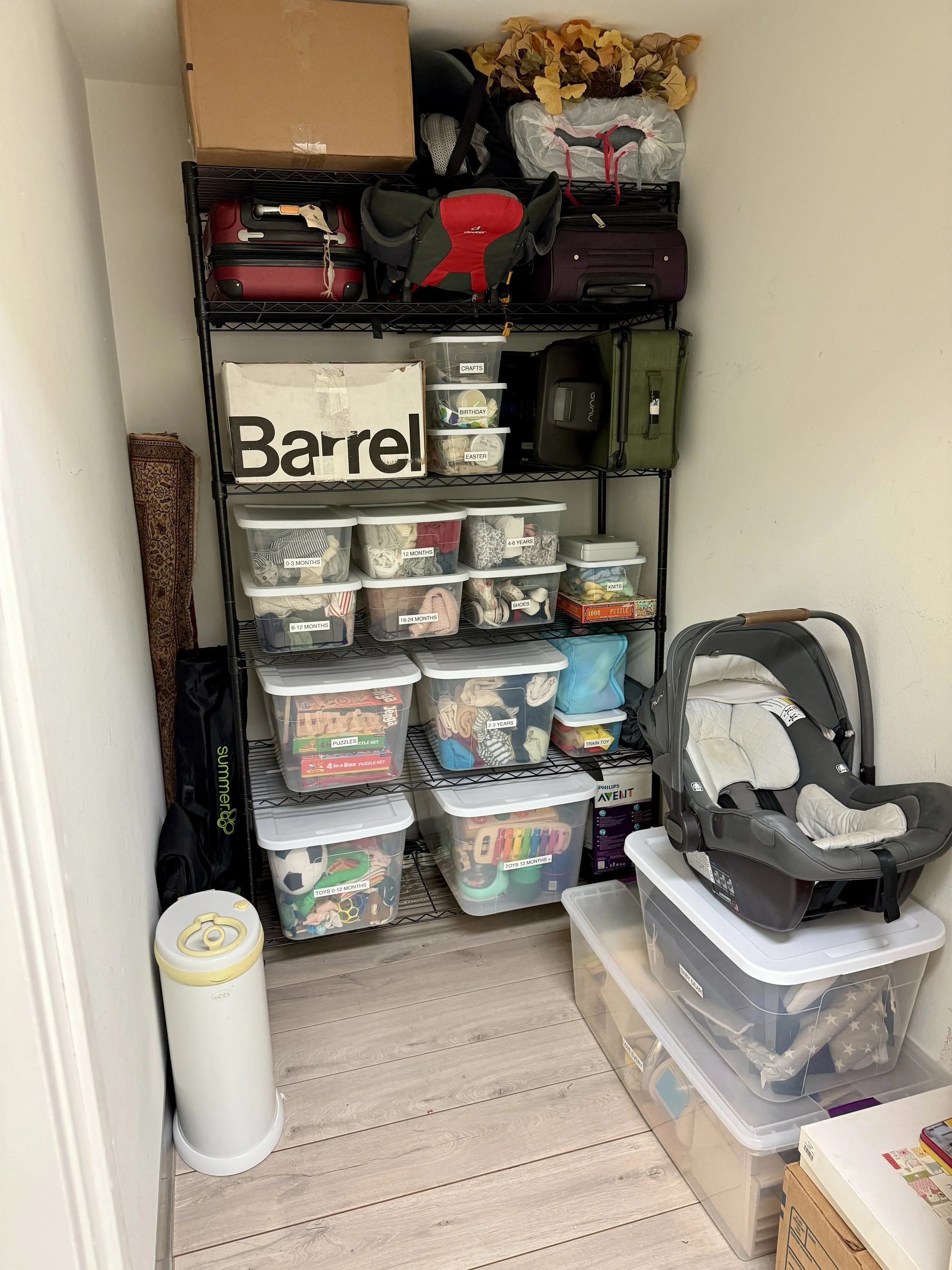 Small storage closet filled with plastic bins holding toys, clothing, and miscellaneous items. Includes a baby car seat, boxed baby swing, shelf with travel and diaper bags, and a humidifier on the floor. Styled by Tidy Doon, Los Angeles.
