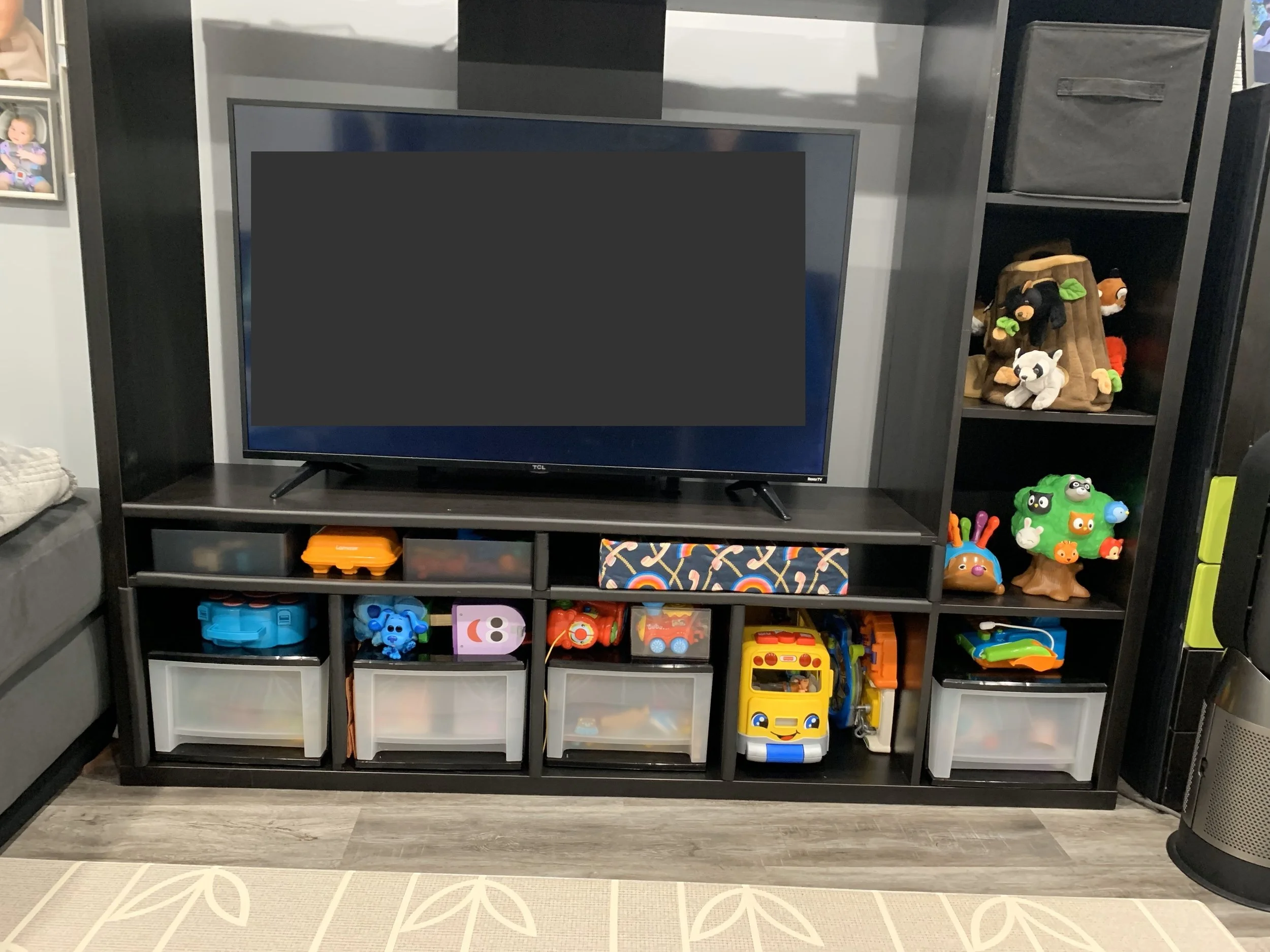 Black entertainment center with a TV on top, toy storage bins, and shelves holding plush toys including a stuffed tree, raccoon, and bear. Various toys and items scattered on the floor. Styled by Tidy Doon, Los Angeles.