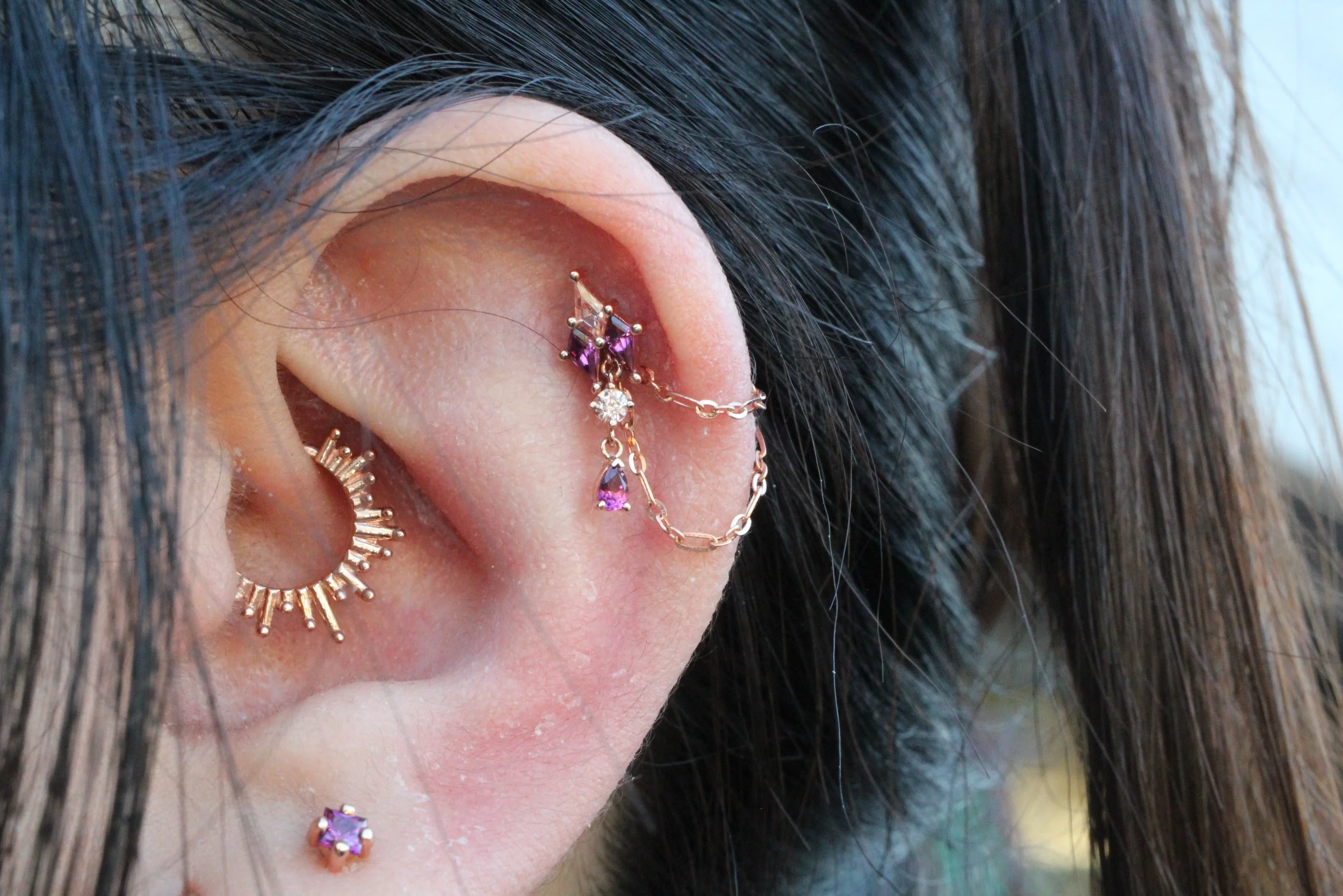 Close-up of a person's ear with multiple piercings, including a gold hoop with hanging purple and clear gemstones, a small purple gemstone stud, gold hoop with projections, and a small stud with a purple gemstone. The person has dark hair partially covering the ear.