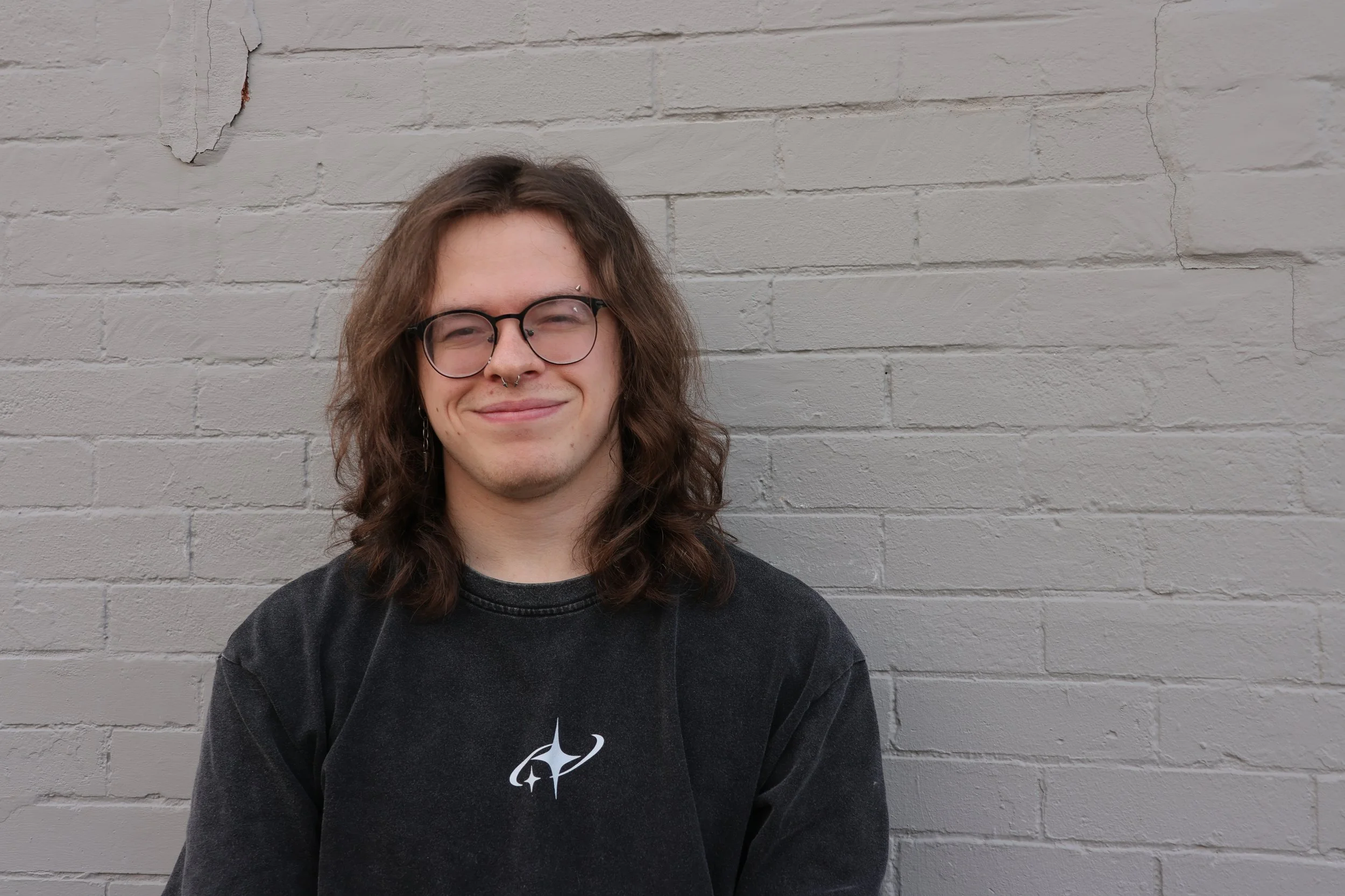 Young man with shoulder-length brown hair, glasses, and a septum piercing, wearing a t-shirt standing against a white brick wall with some peeling paint, posing 