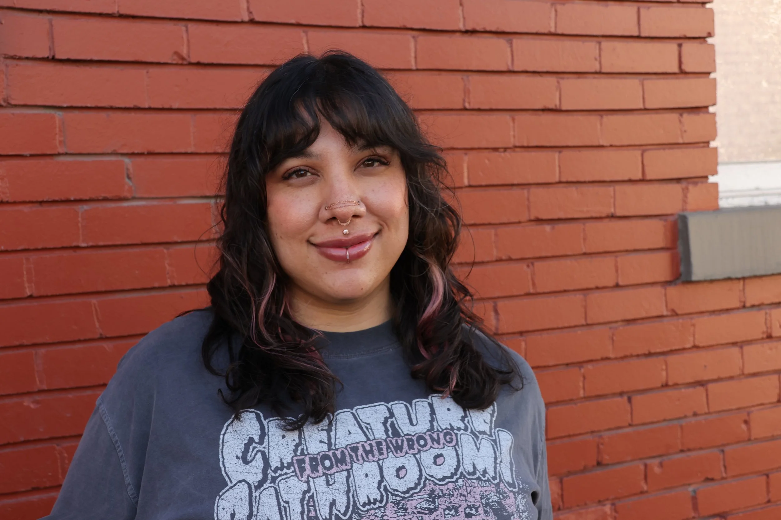 Young woman standing against a red brick wall, wearing a t-shirt and pink highlights in her dark hair, smiling at the camera.