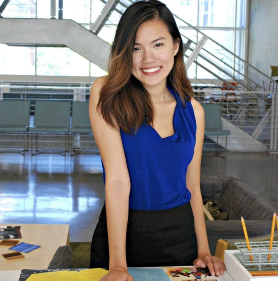 A young woman with shoulder-length brown hair smiling, wearing a blue sleeveless top, standing at a table in a modern, glass-filled building with stairs in the background.