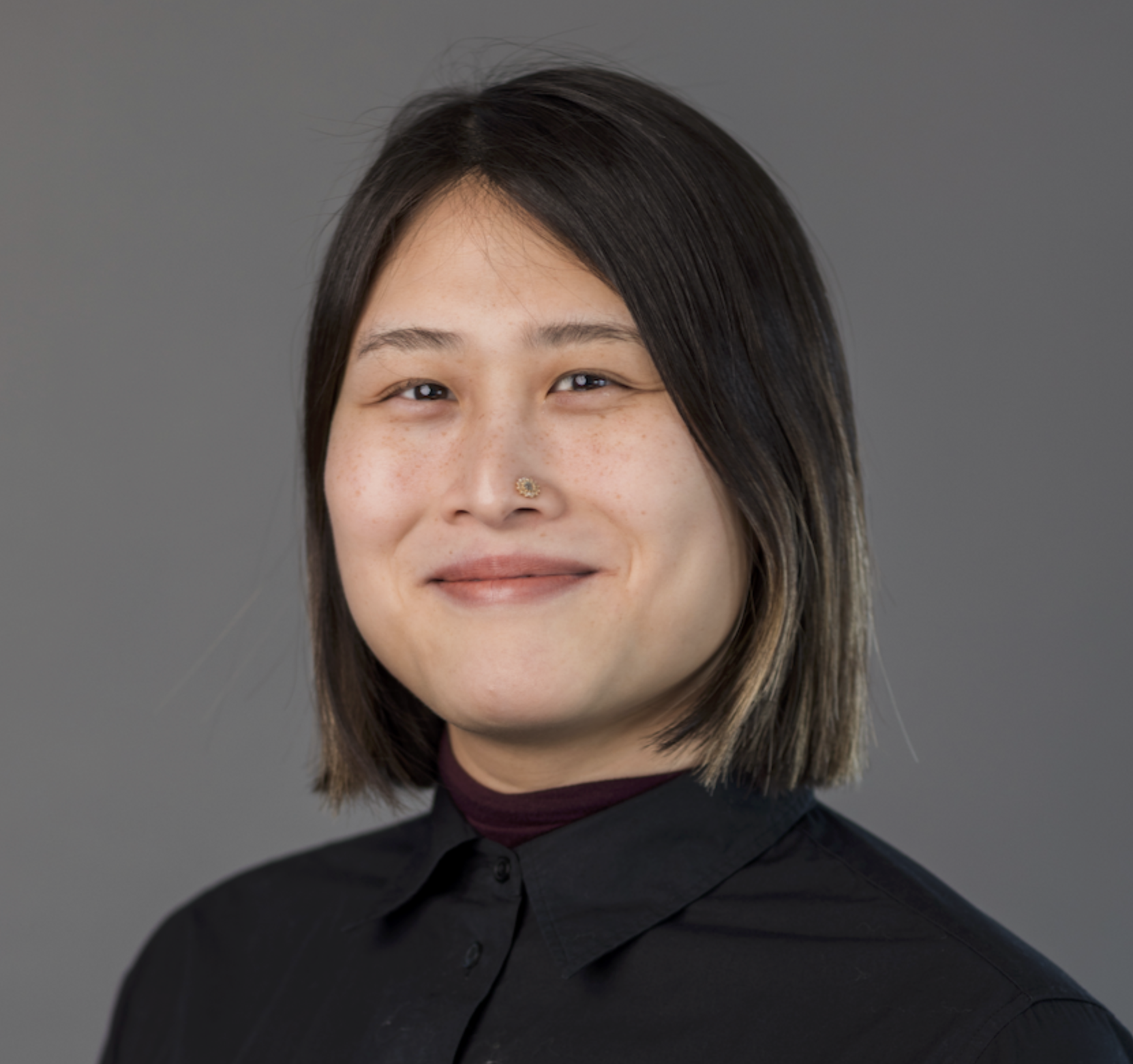 Portrait of a woman with shoulder-length dark brown hair with highlights, wearing a black shirt and a maroon turtleneck, standing against a plain gray background.