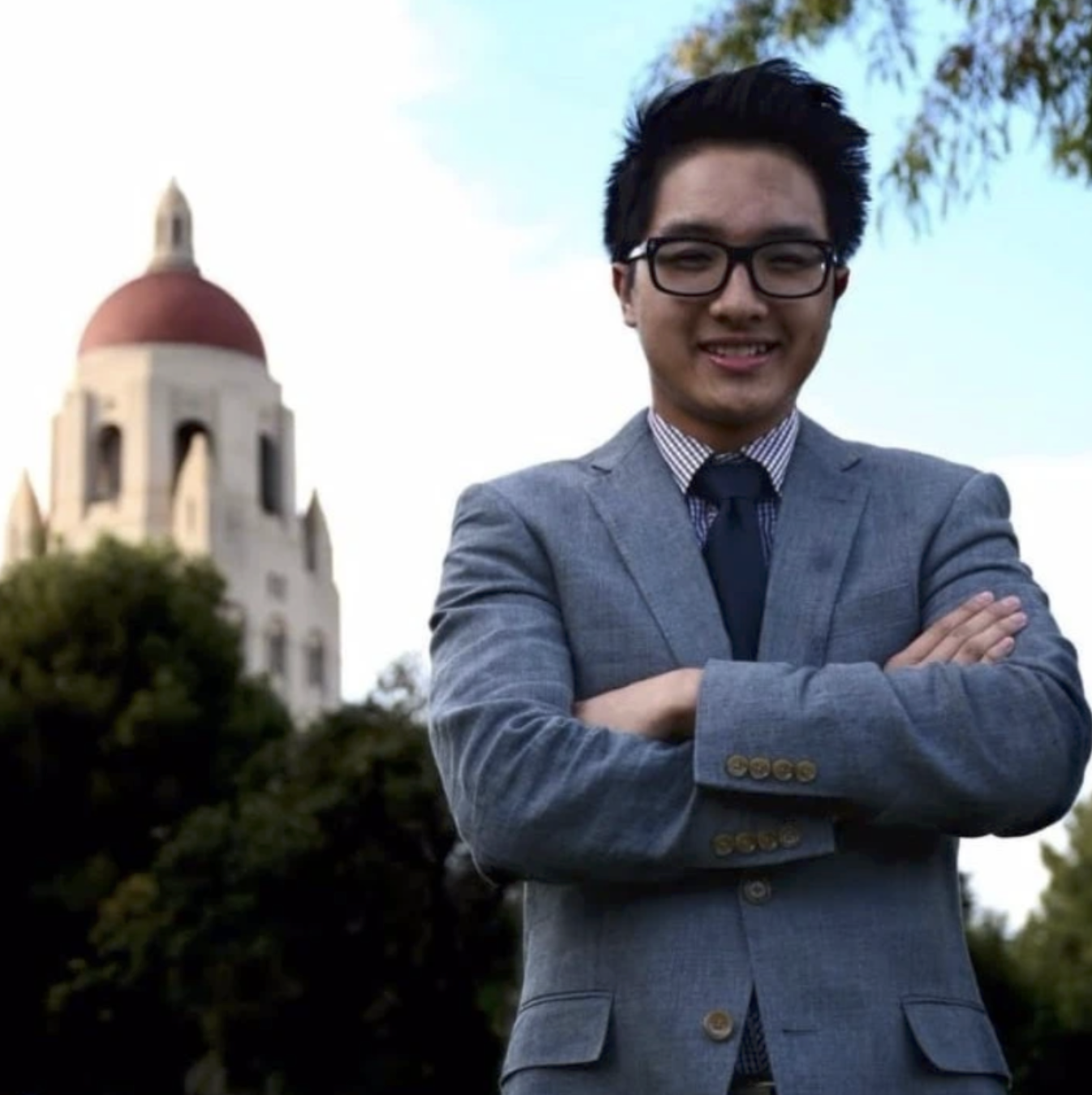 A man in a gray suit with a striped shirt and dark tie standing outdoors with arms crossed, smiling. In the background, there is a building with a red dome and a steeple, surrounded by trees and bushes, with a blue sky.