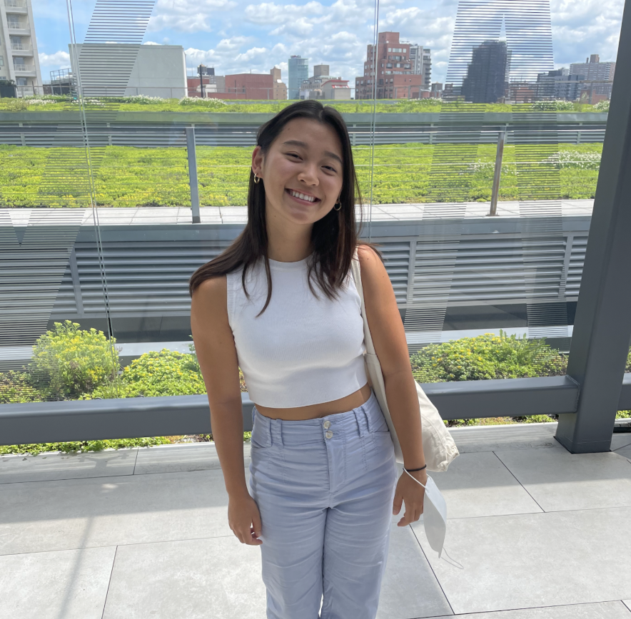 A young woman smiling and standing outdoors in front of a glass wall with a city skyline in the background. She is wearing a white sleeveless crop top and light-colored pants, and holding a face mask in her hand.