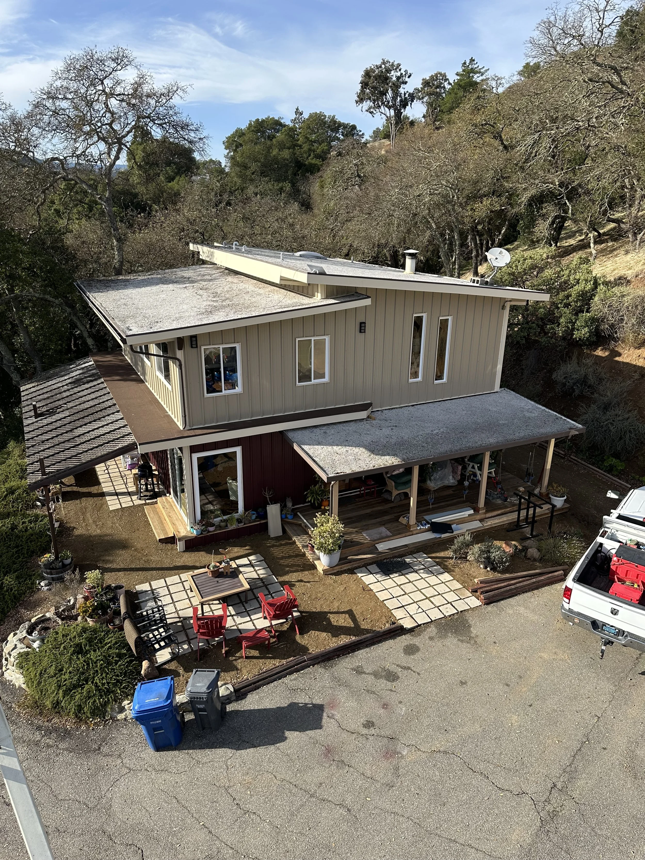 A modern two-story house with a beige upper floor and red lower floor, surrounded by a small outdoor patio with red chairs, a table, and potted plants, set in a natural, wooded area with trees and a gravel driveway.
