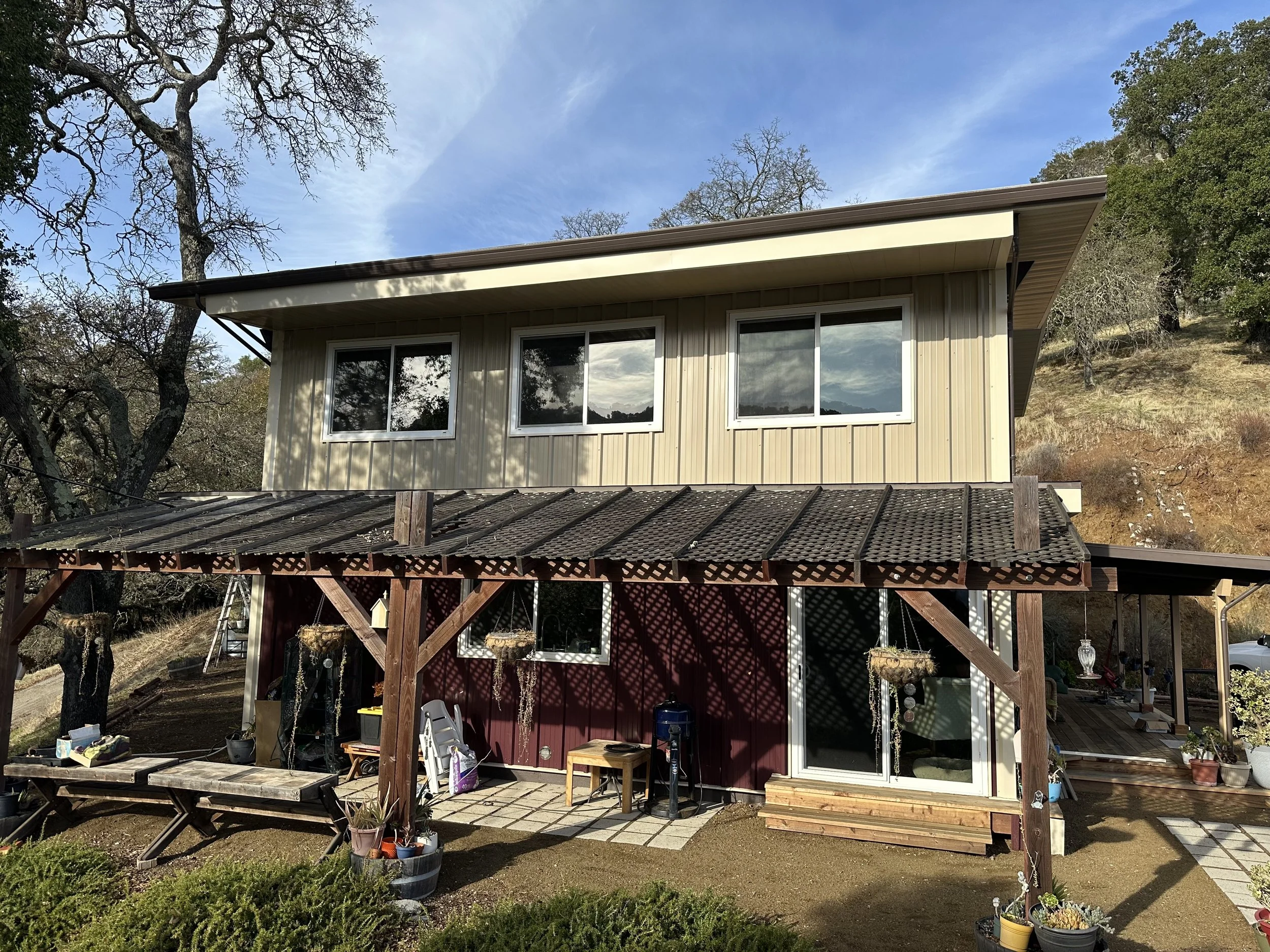 Two-story house with beige siding on upper level and red lower level, large windows on upper floor, covered porch with wooden beams and lattice roof, potted plants, outdoor furniture, and various garden decorations.