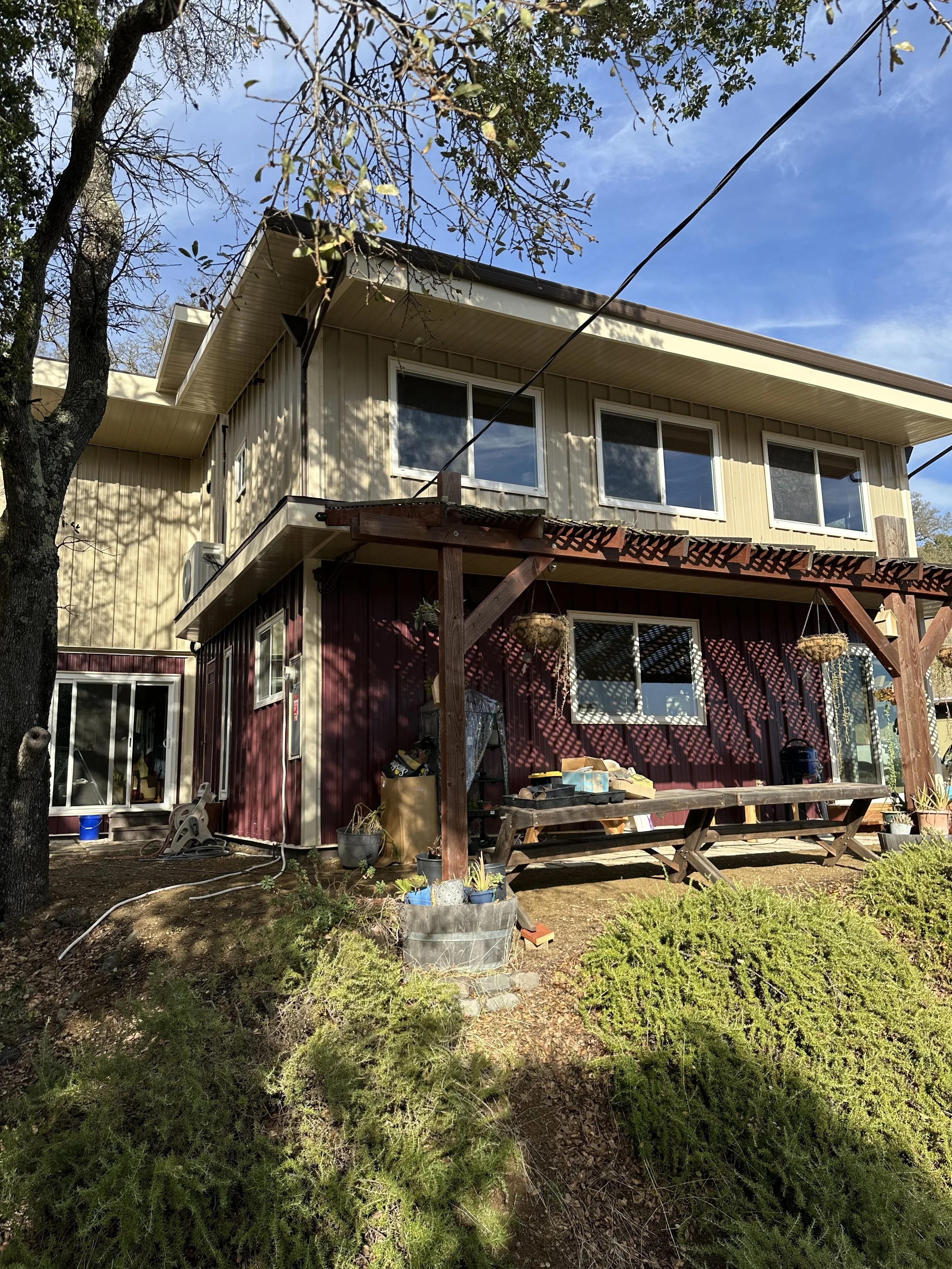A two-story house with beige upper siding and red lower siding, featuring large windows on the second floor and a sliding glass door on the ground level. There is a wooden pergola over a bench on the patio. The yard has bushes, potted plants, and gardening tools, and the scene is lit by sunlight with a blue sky overhead.