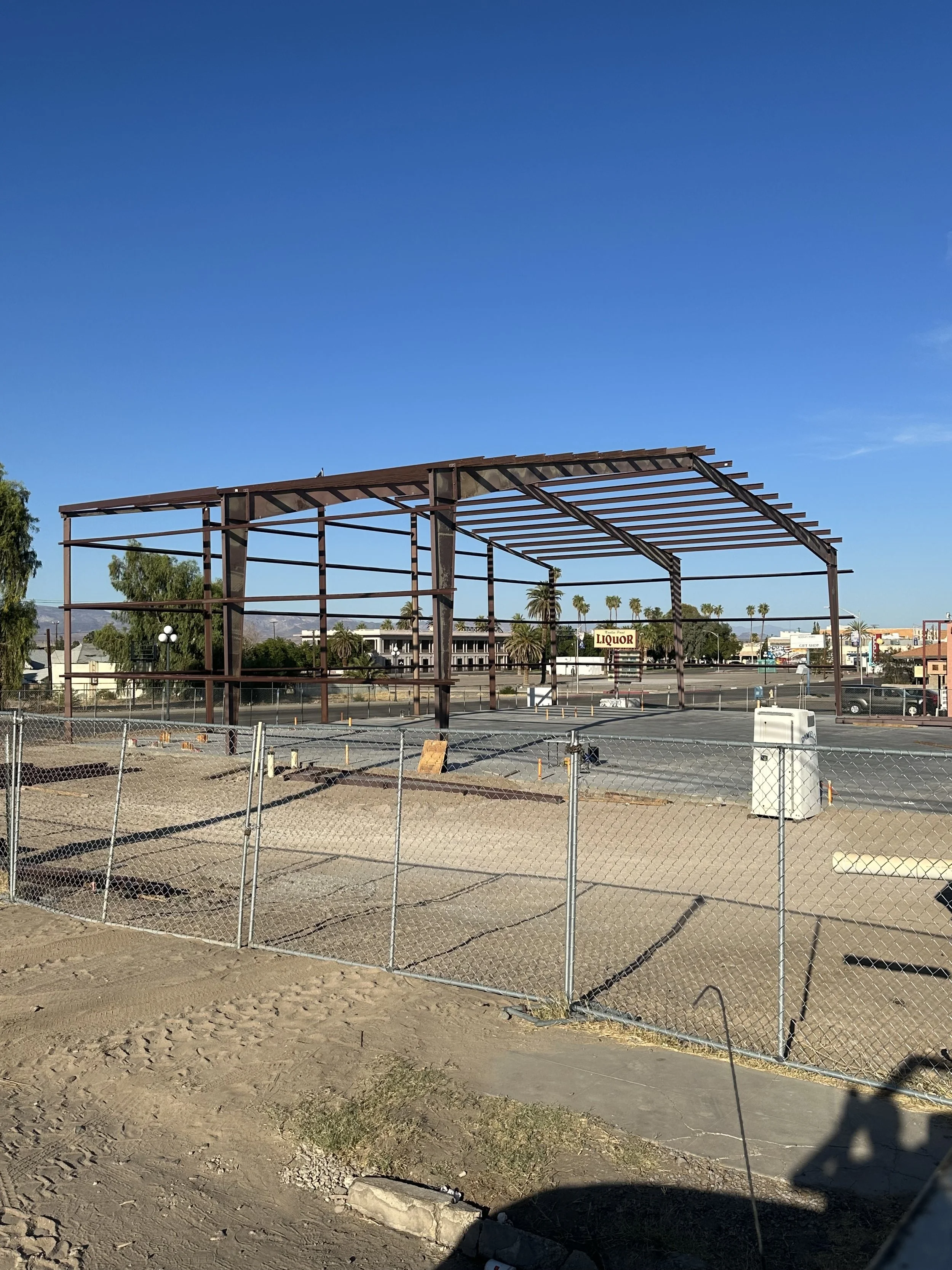 Sidewalk view of a metal framing to a steel building. Wire fencing around the lot with a concrete slab base