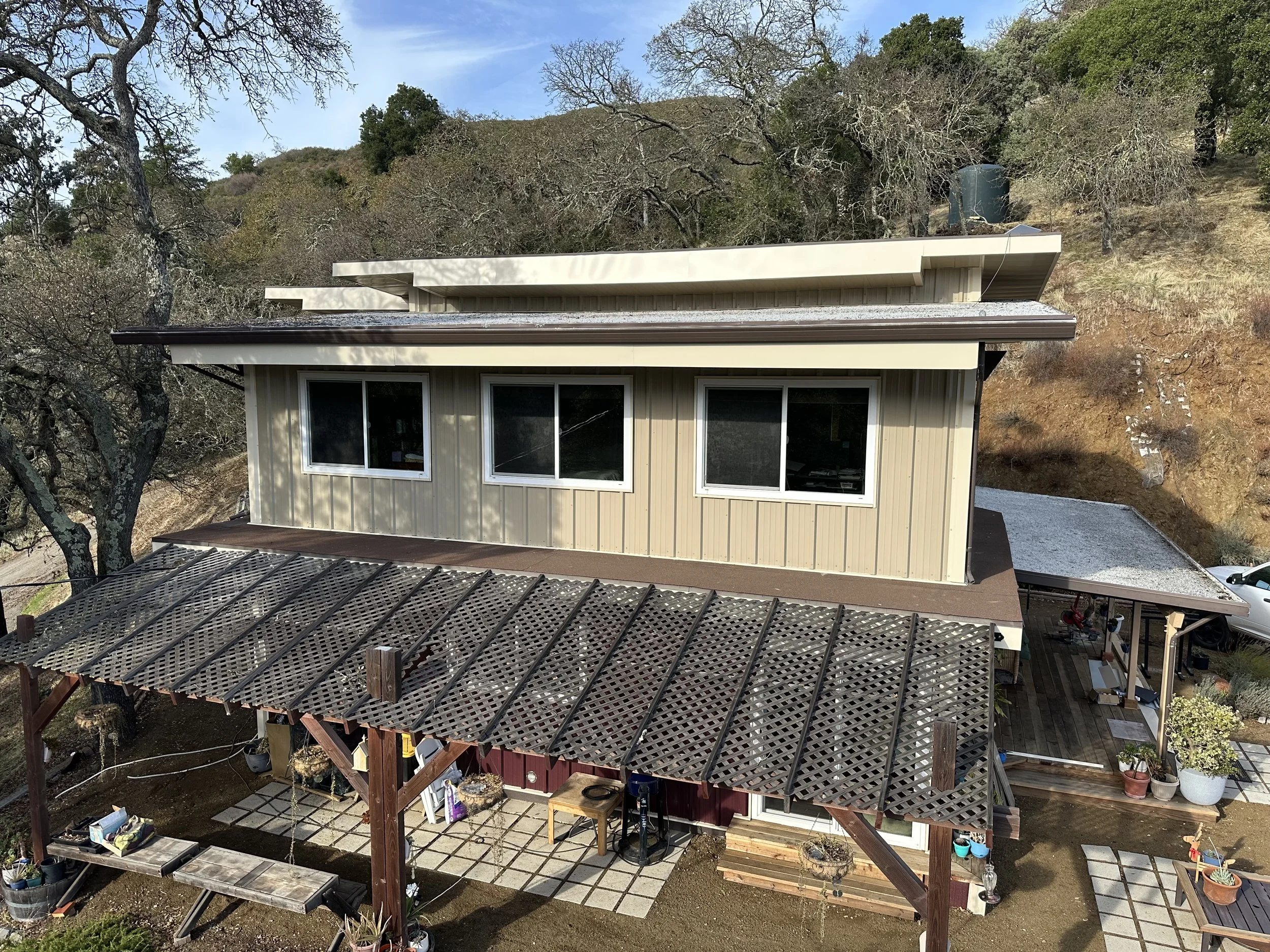 A two-story house with beige siding, three large windows on the upper floor, a brown roof, and a covered porch with a lattice roof. The house is situated on a hillside with trees around, and there is a gravel area on the right side with a vehicle parked nearby.