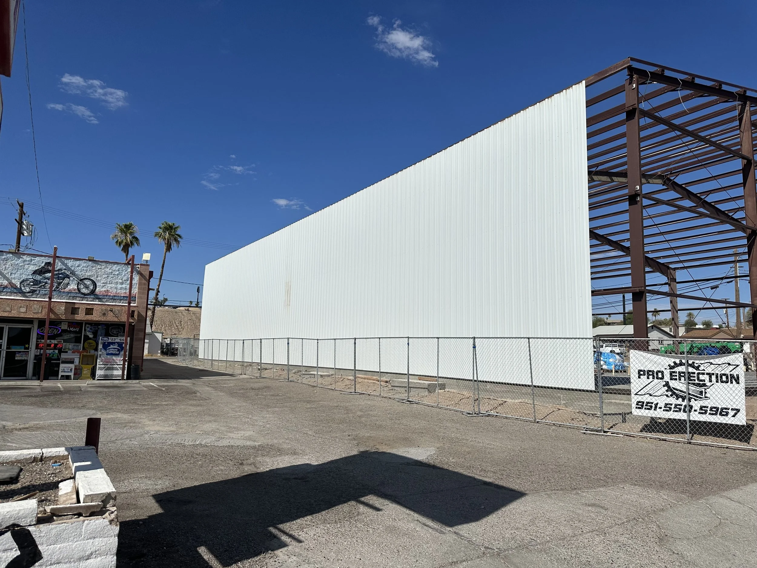 Back Back view of the steel building with siding added and framing of the roof showing. Pro Erection banner with phone number on a wire fencing. 