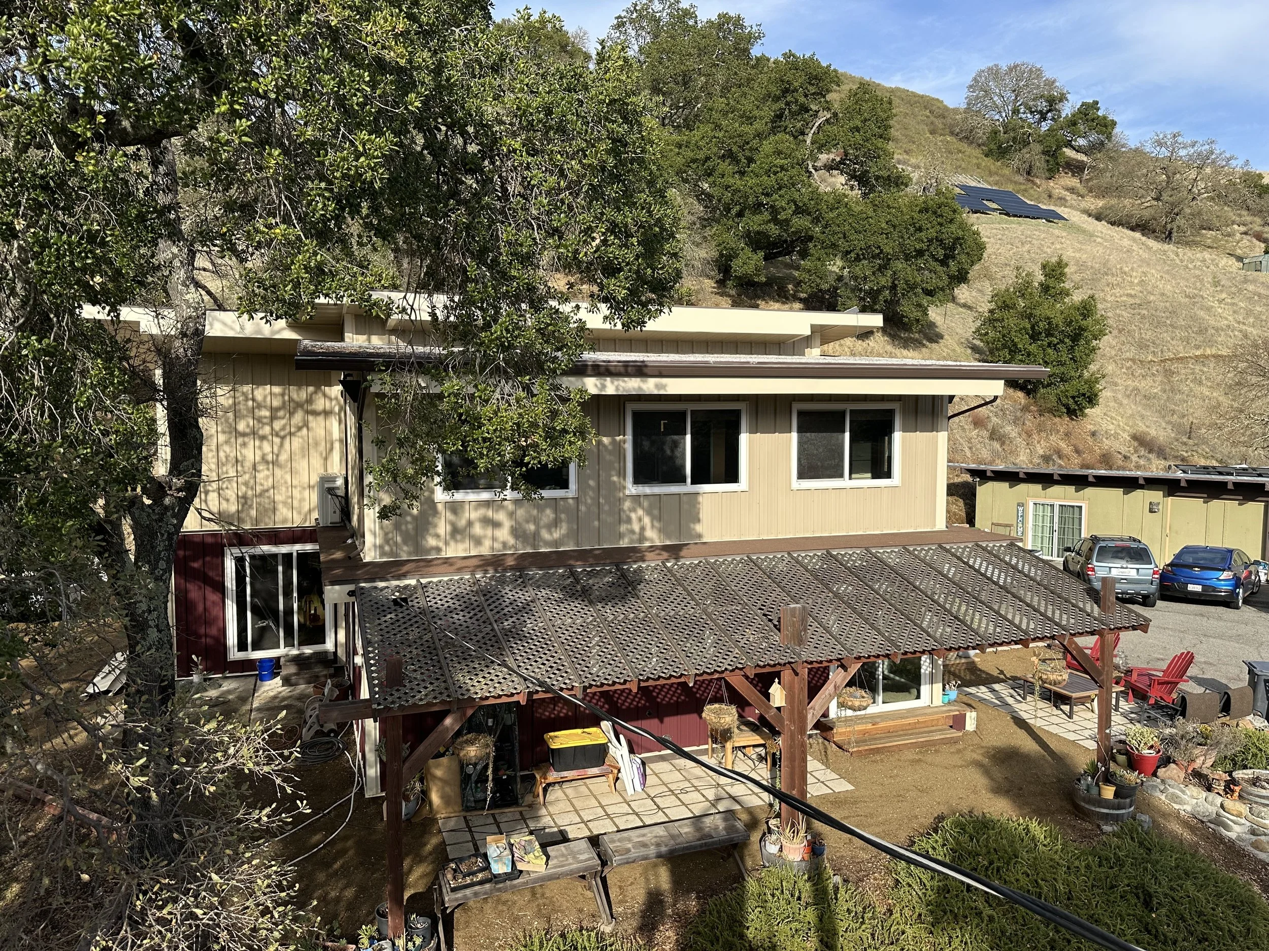 Residential house with beige siding, large windows, and a covered patio with a lattice roof. There are trees, a driveway with parked cars, and a hillside in the background.