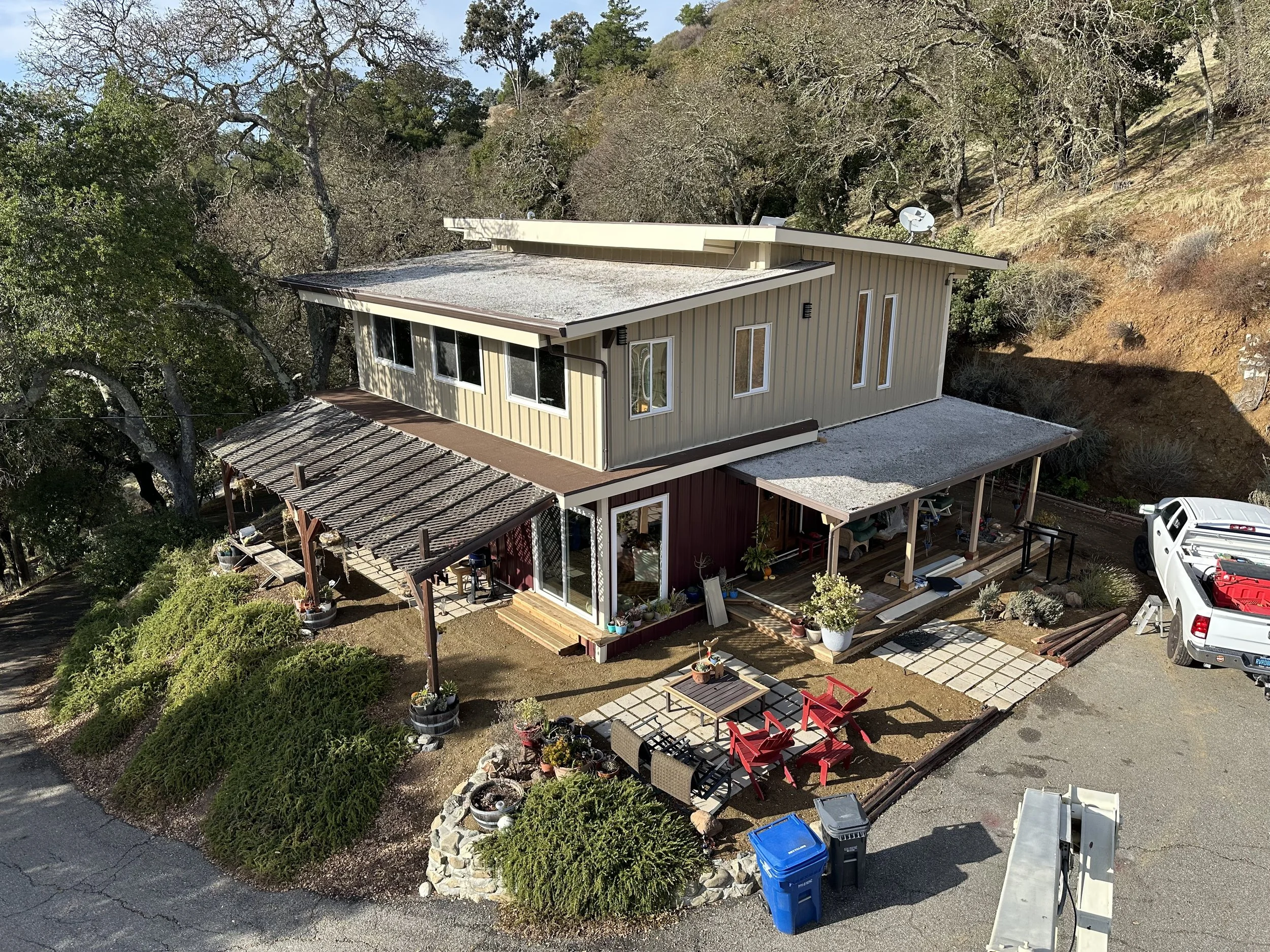 An aerial view of a two-story house with a mix of modern and rustic design, surrounded by trees and a dirt hillside. The house features beige vertical siding, a metal roof, and a covered porch with outdoor furniture. There is a paved seating area with red chairs and a table, a gravel driveway, and a white truck parked nearby.