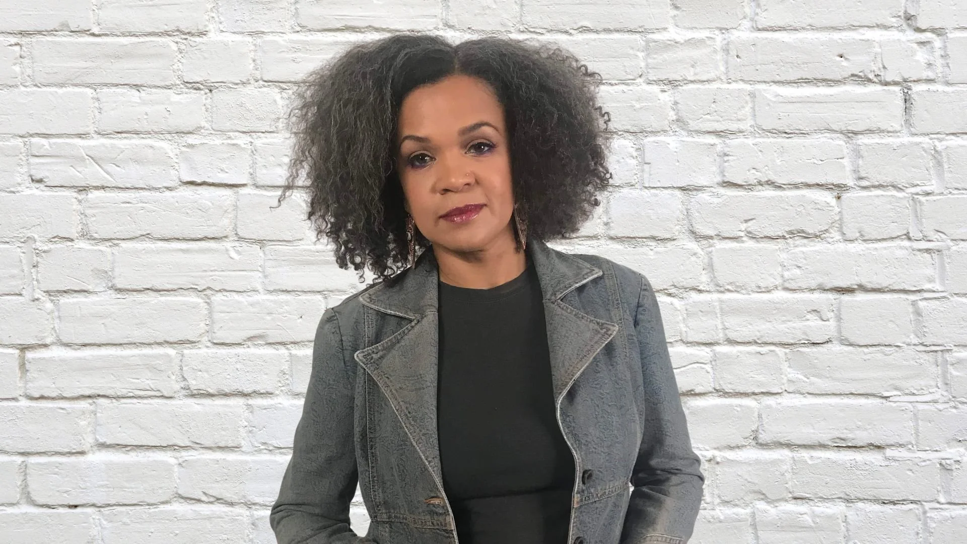 Portrait of Kim Wilson, Ph.D., with dark curly hair, wearing a denim blazer over a black top, in front of a white brick wall.