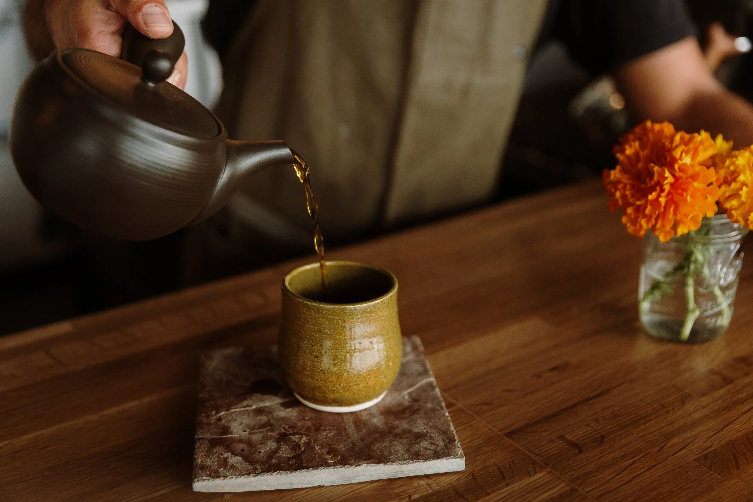 Pouring tea into a ceramic cup on a wooden table, with a small jar of orange flowers nearby.