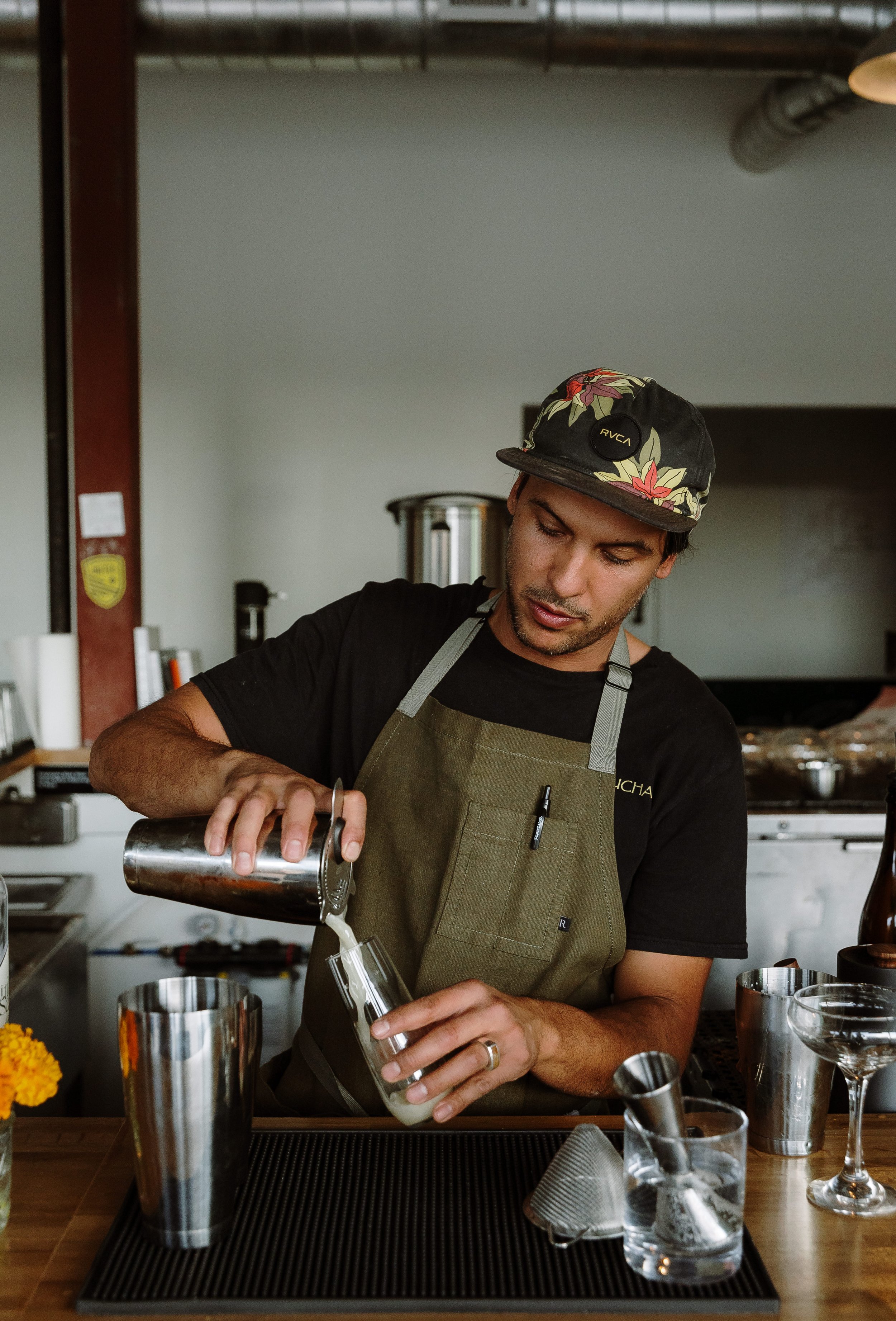 A bartender wearing a cap and apron prepares a cocktail at a bar, pouring a liquid from a shaker into a glass.