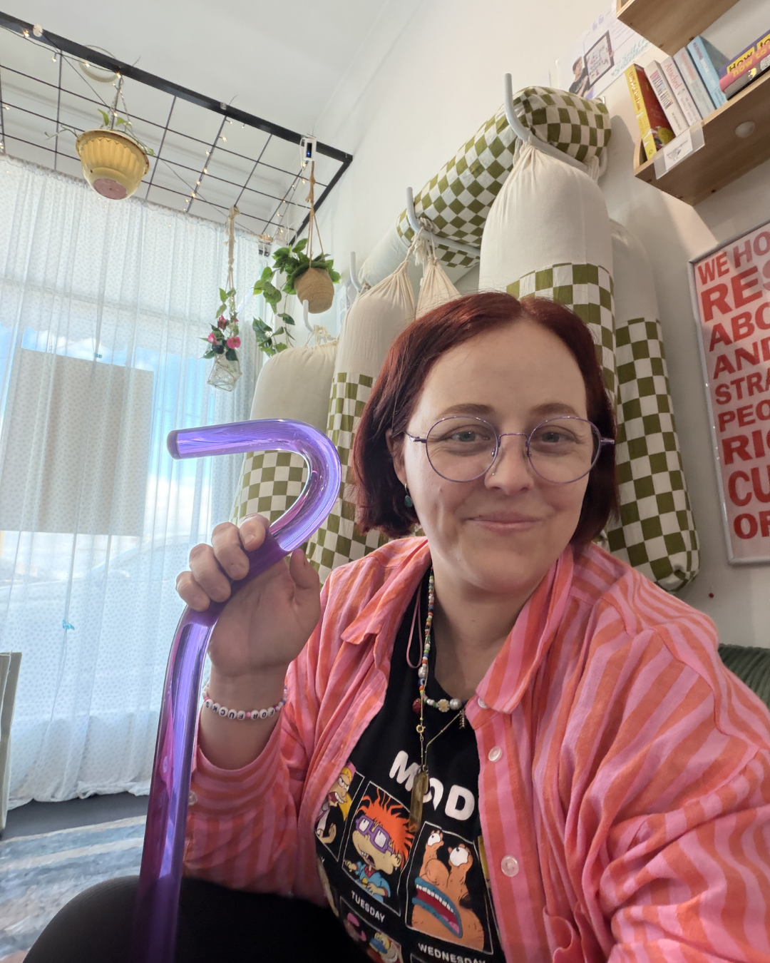 A woman with short red hair, wearing glasses, a black graphic t-shirt, and a pink striped shirt, smiling while holding a purple light-up cane in a room decorated with hanging plants, a white curtain, and colorful posters.