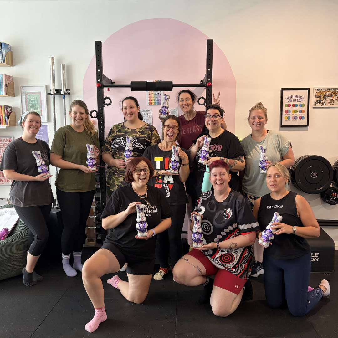 Group of ten women in a gym holding purple bunny-shaped chocolate or cake treats, smiling for a photo in front of a pink wall and fitness equipment.