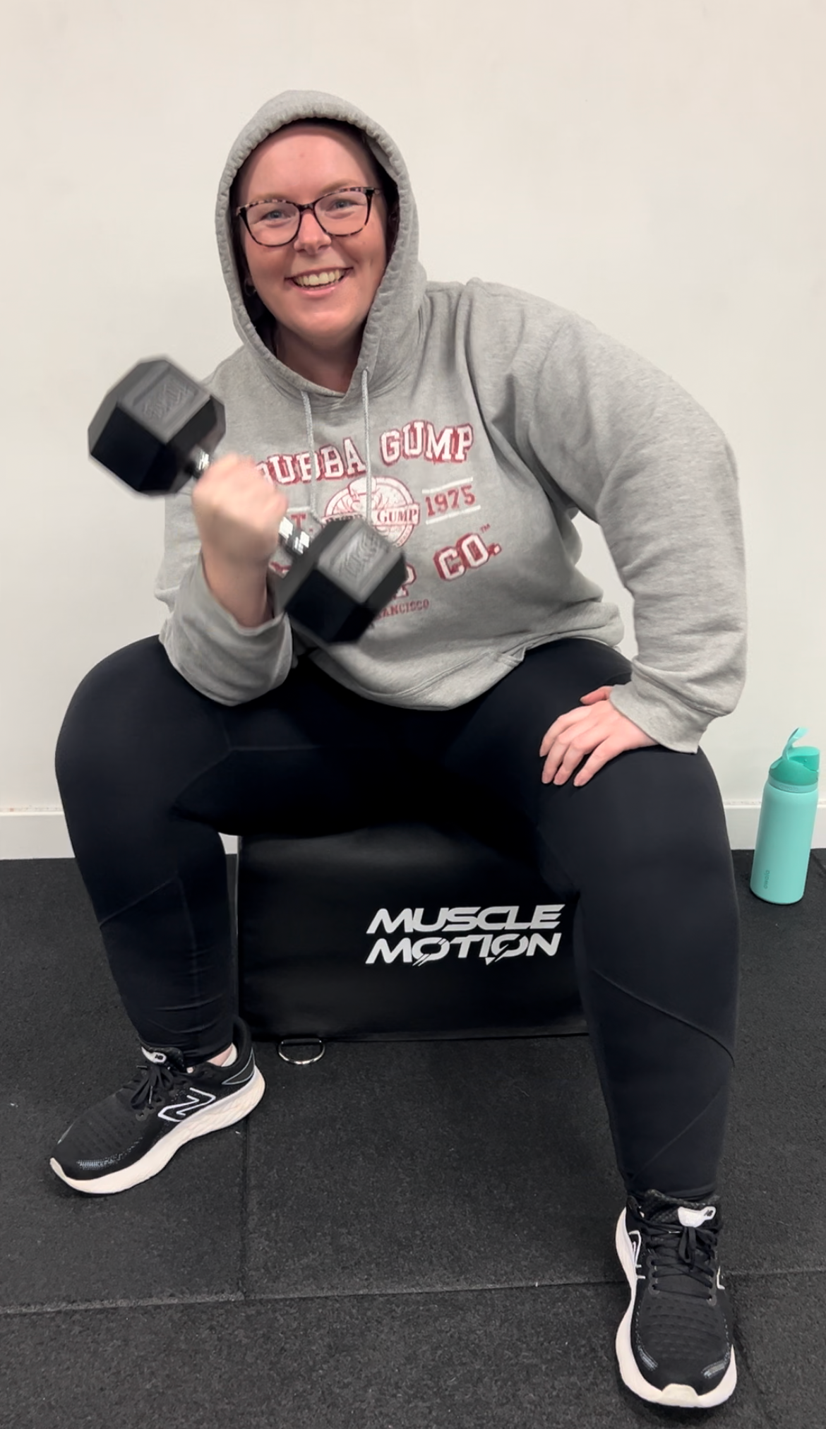 A woman in a gray hoodie, glasses, and black leggings, smiling while lifting a black dumbbell in a fitness gym. She is sitting on a black exercise ball labeled 'MUSCLE MOTION,' with a water bottle on the floor nearby.