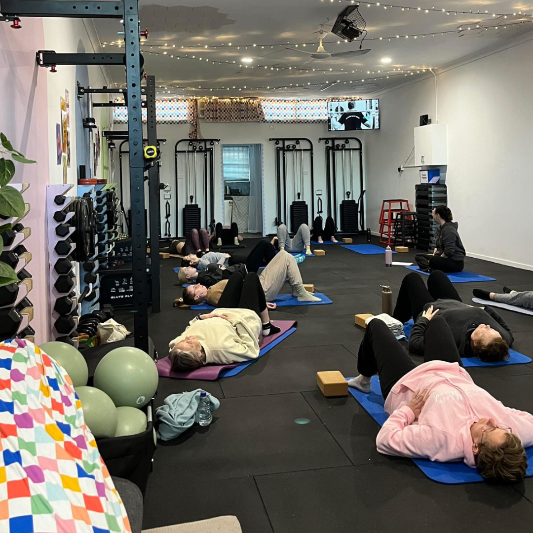 A group fitness class with participants lying on mats in a gym, performing stretches or exercises under a ceiling decorated with string lights, with fitness equipment and a small TV in the background.