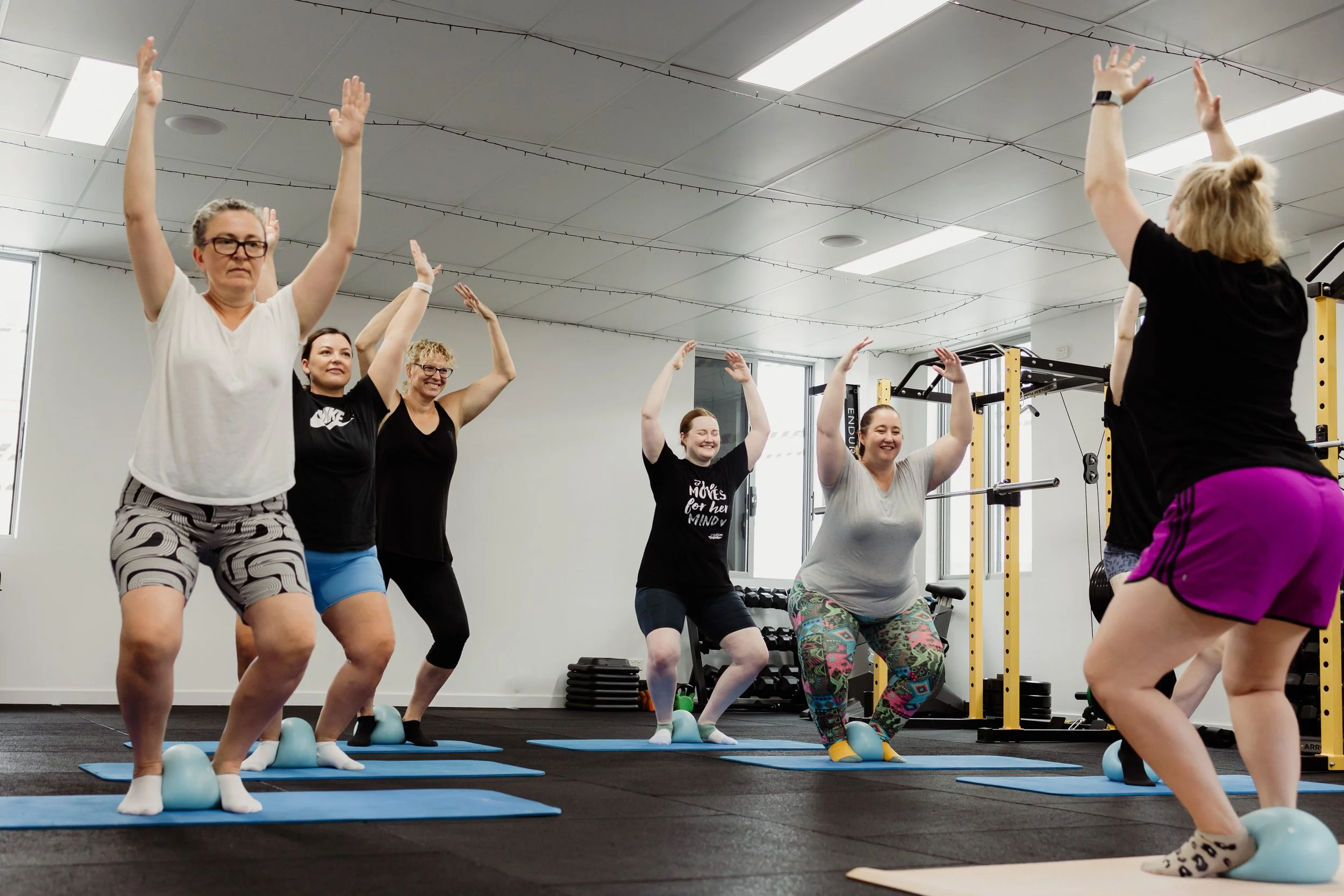 Group of women participating in a fitness class, practicing yoga or balance exercises on blue mats with small workout balls in a gym or fitness studio.