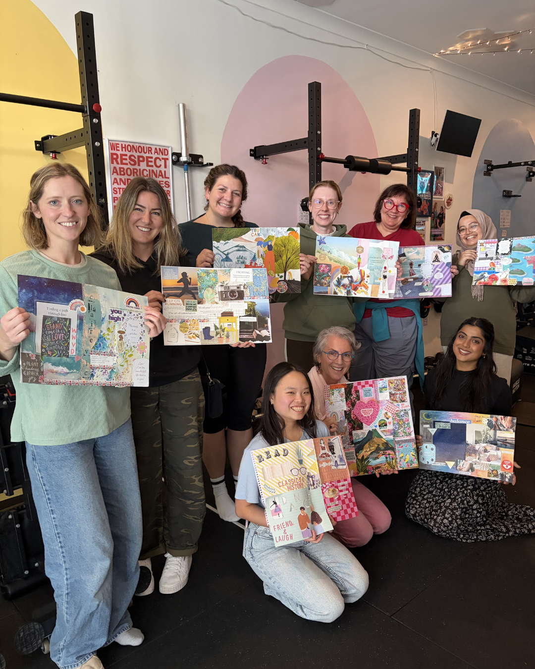 A group of ten women and girls smiling and holding colorful scrapbook pages in a gym or fitness room with workout equipment and a sign on the wall.