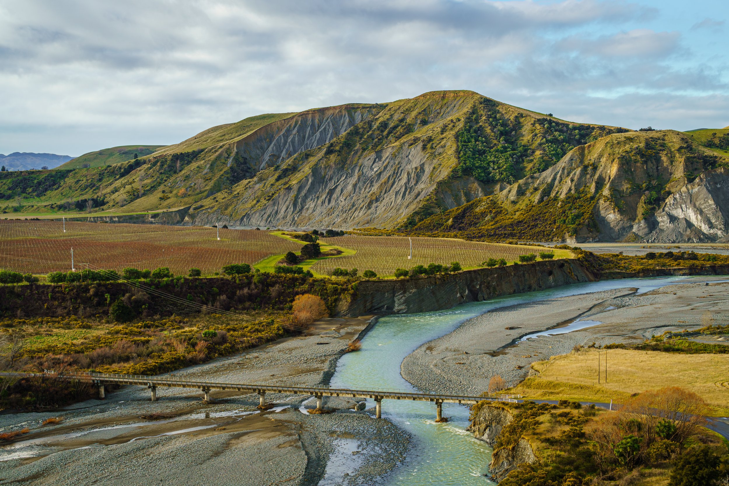 Turquoise Awatere River channel flowing under a concrete bridge past vineyard terraces and eroded bluffs in Marlborough